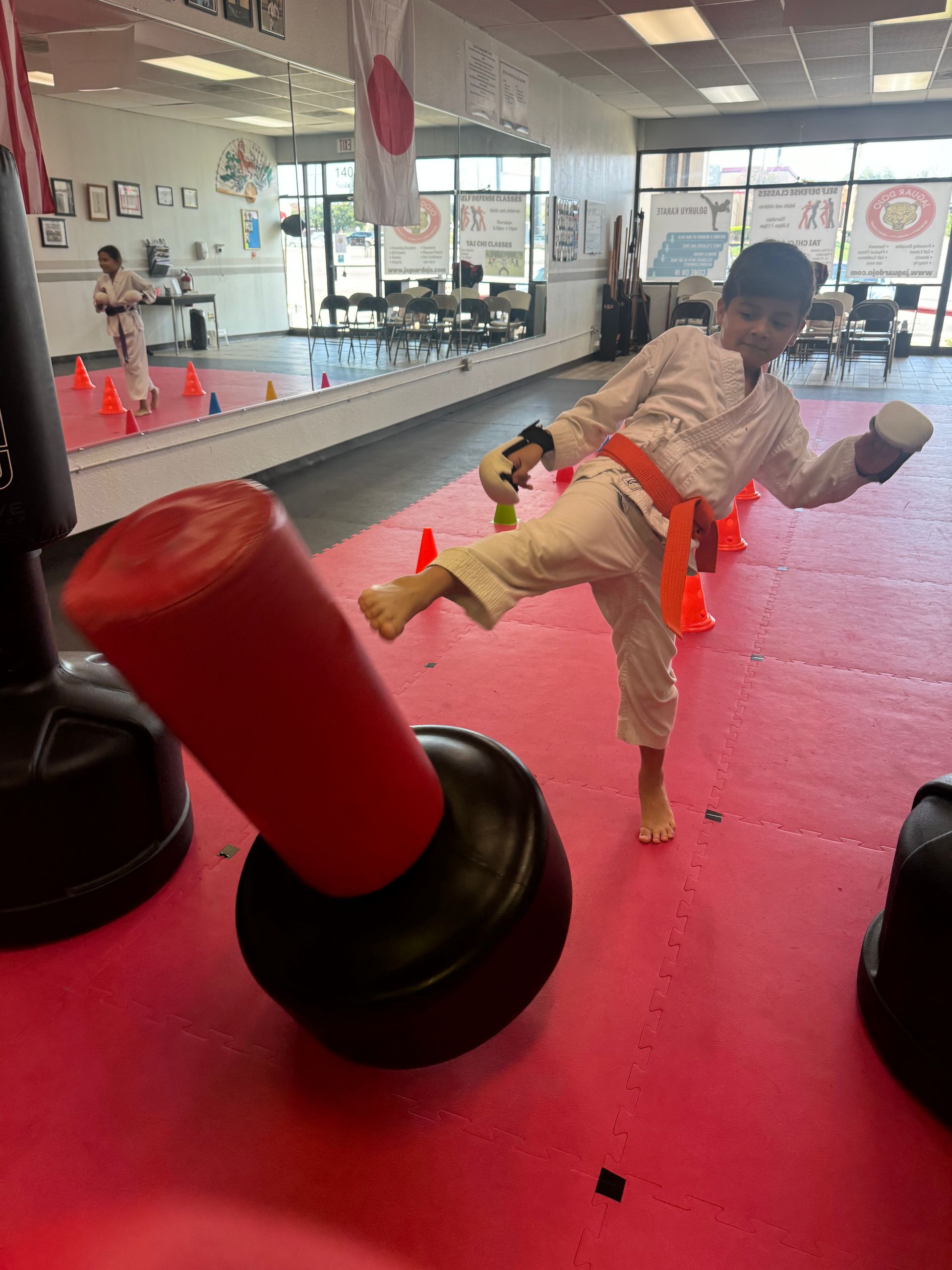 Child kicking a red-and-black punching bag in a martial arts studio with red mats and mirrors