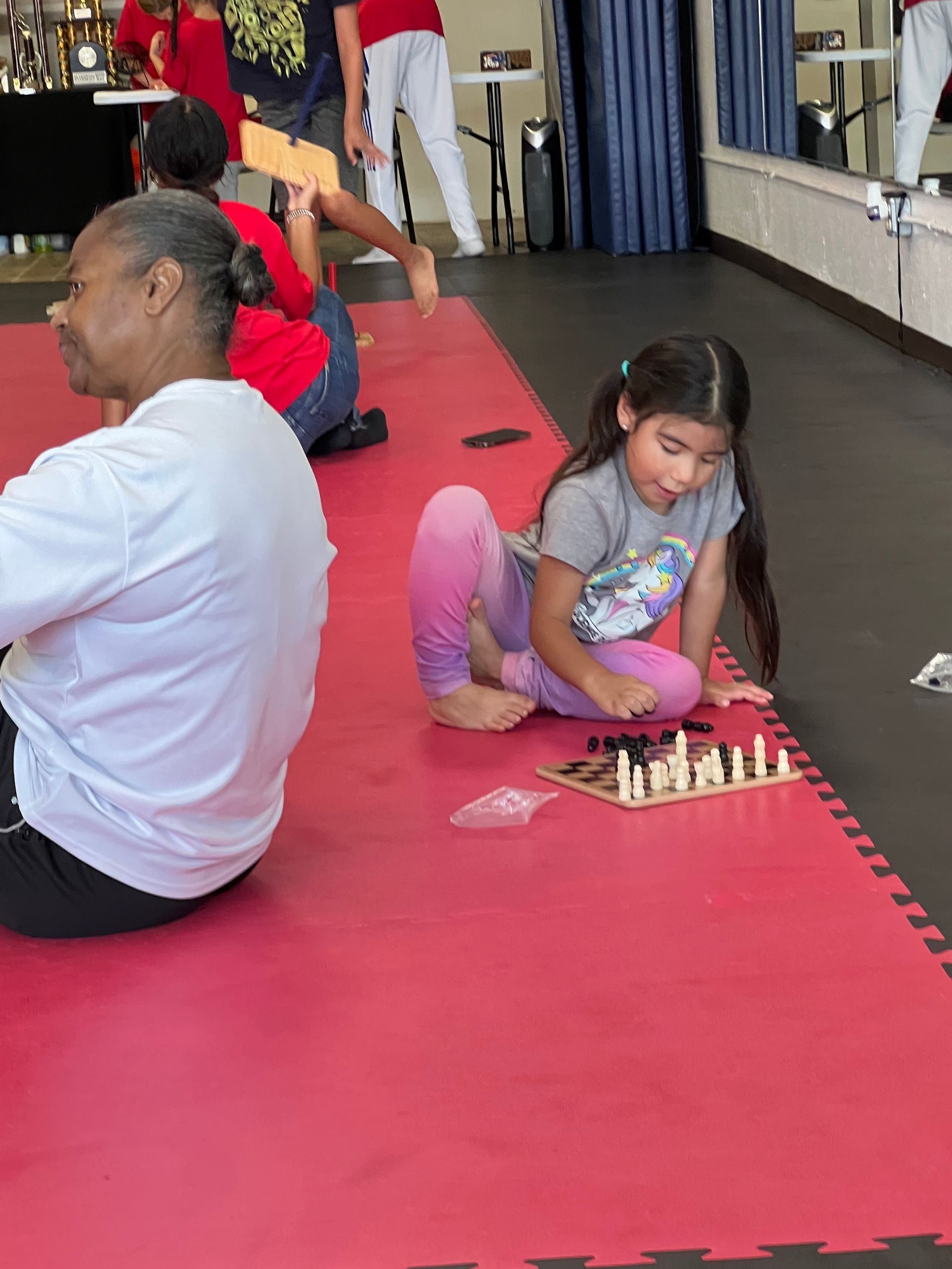 Child sitting on a red mat, playing chess in a bright indoor room