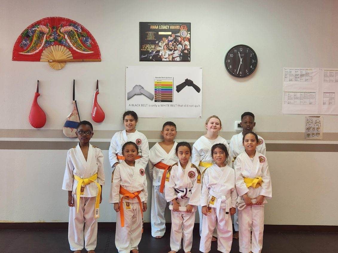 Six children in martial arts uniforms pose in a studio with belts and wall decorations