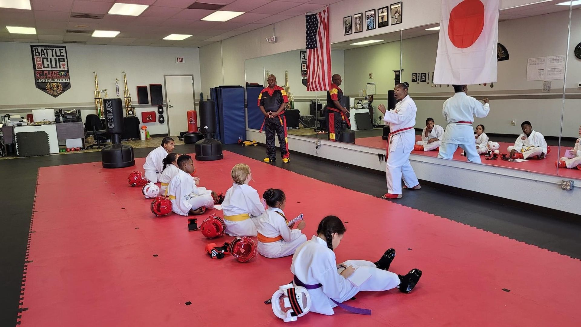 Martial arts class on red mats with students seated and instructor demonstrating near mirrors and flags