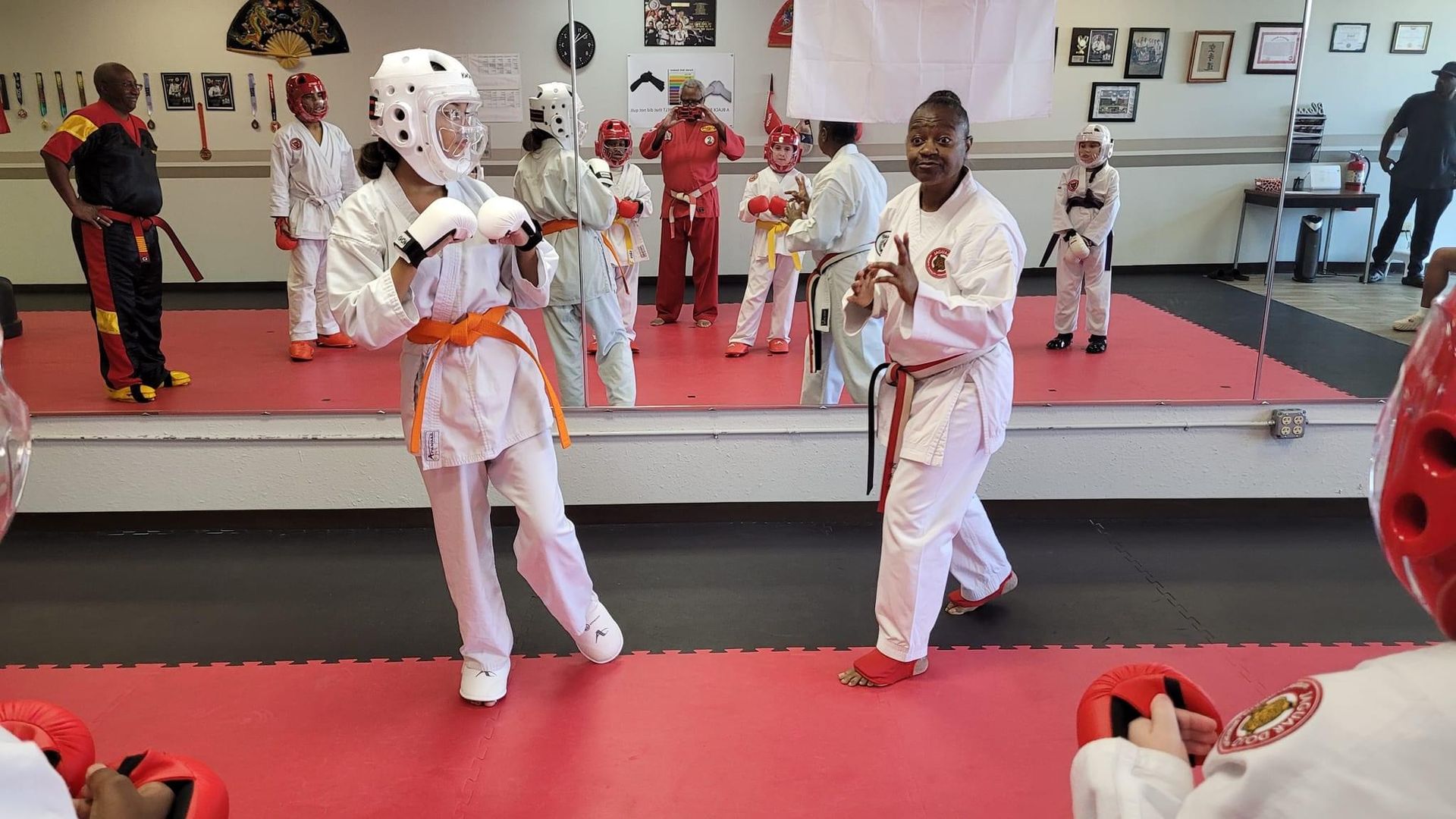 Two martial arts students in white uniforms spar in a dojo with mirrors and red mats.