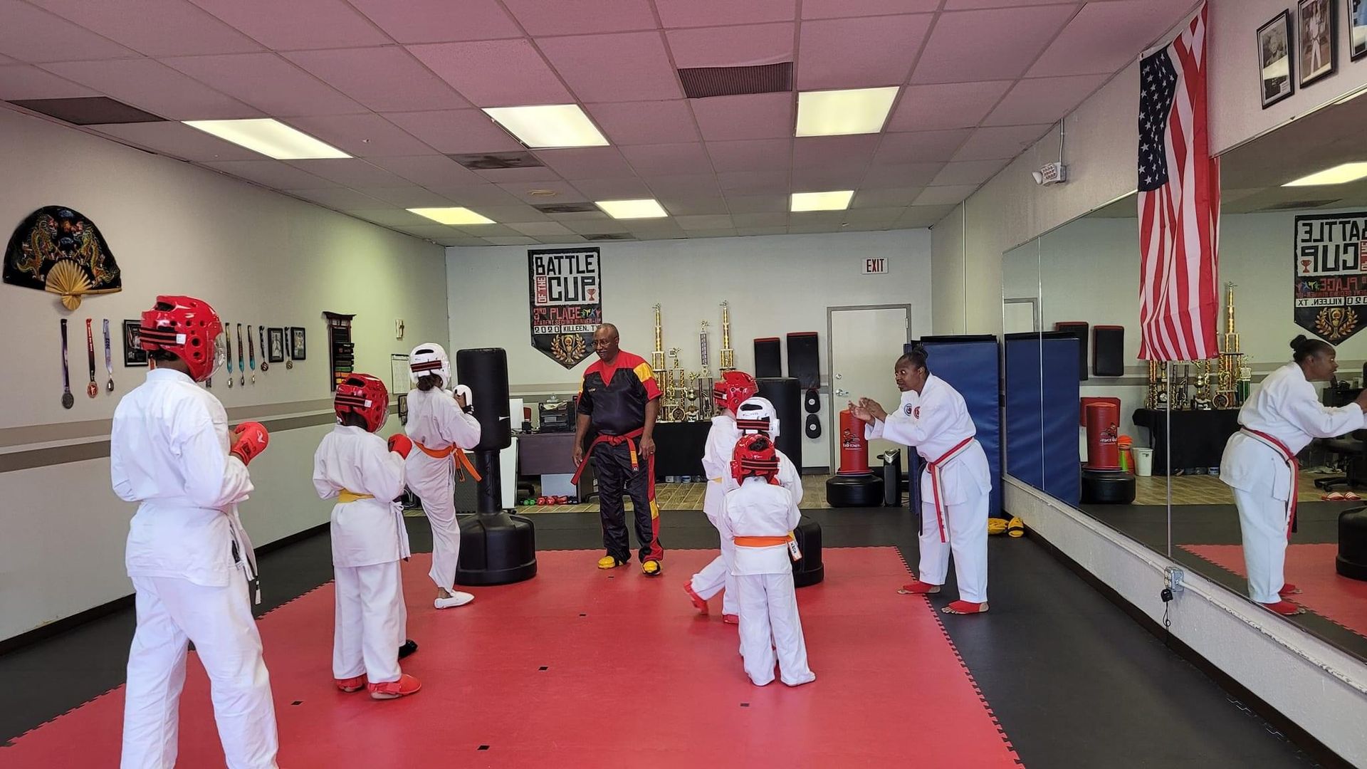 Children in white martial arts uniforms and red headgear practice in a dojo with red mats and mirrors