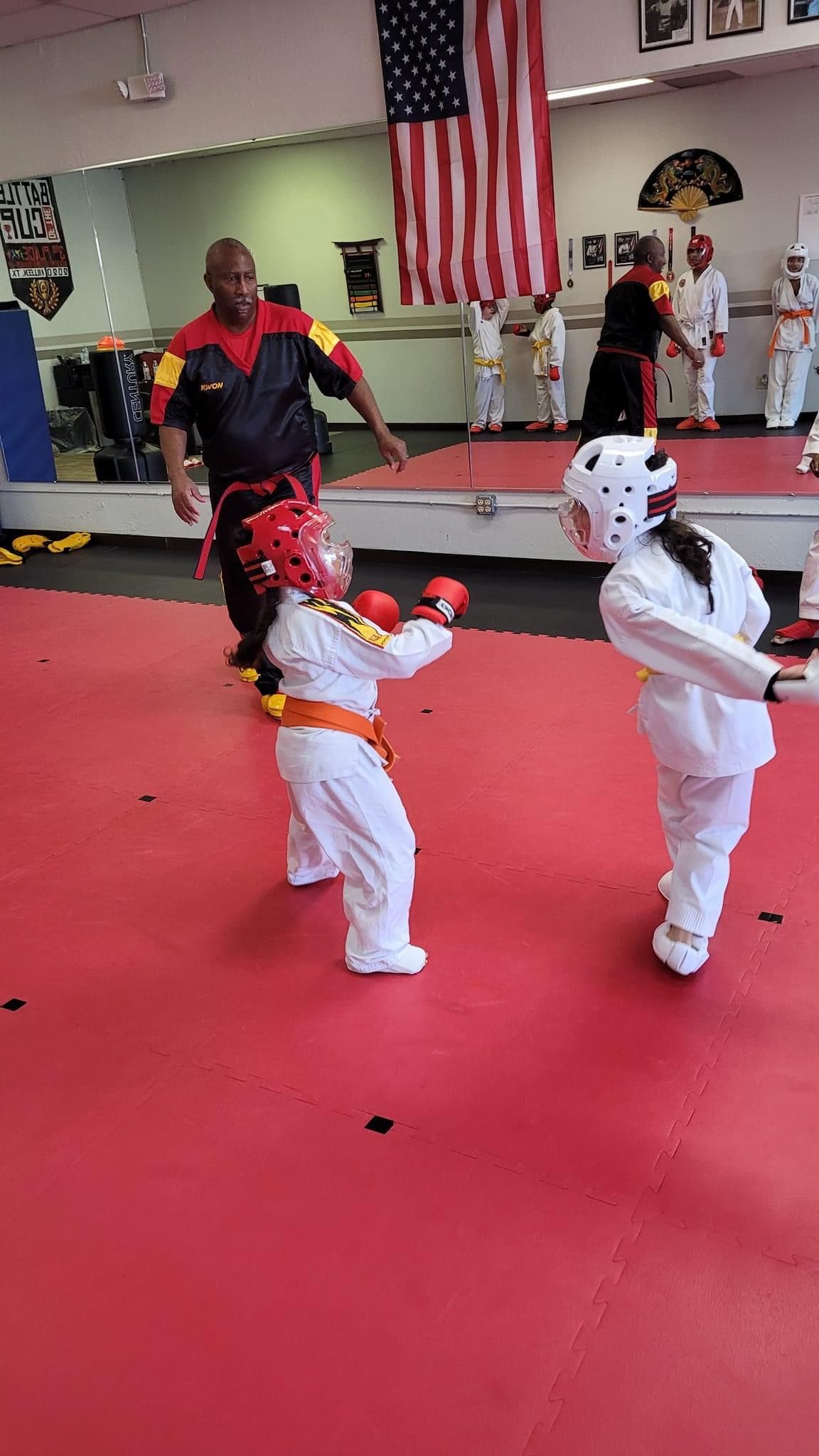 Two children in white martial arts uniforms sparring on a red mat in a dojo, with a coach and American flag behind them.