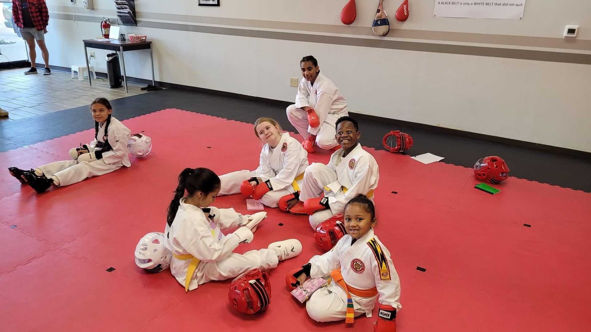 Children in white martial arts uniforms sitting on red mats in a dojo, with helmets and gear nearby