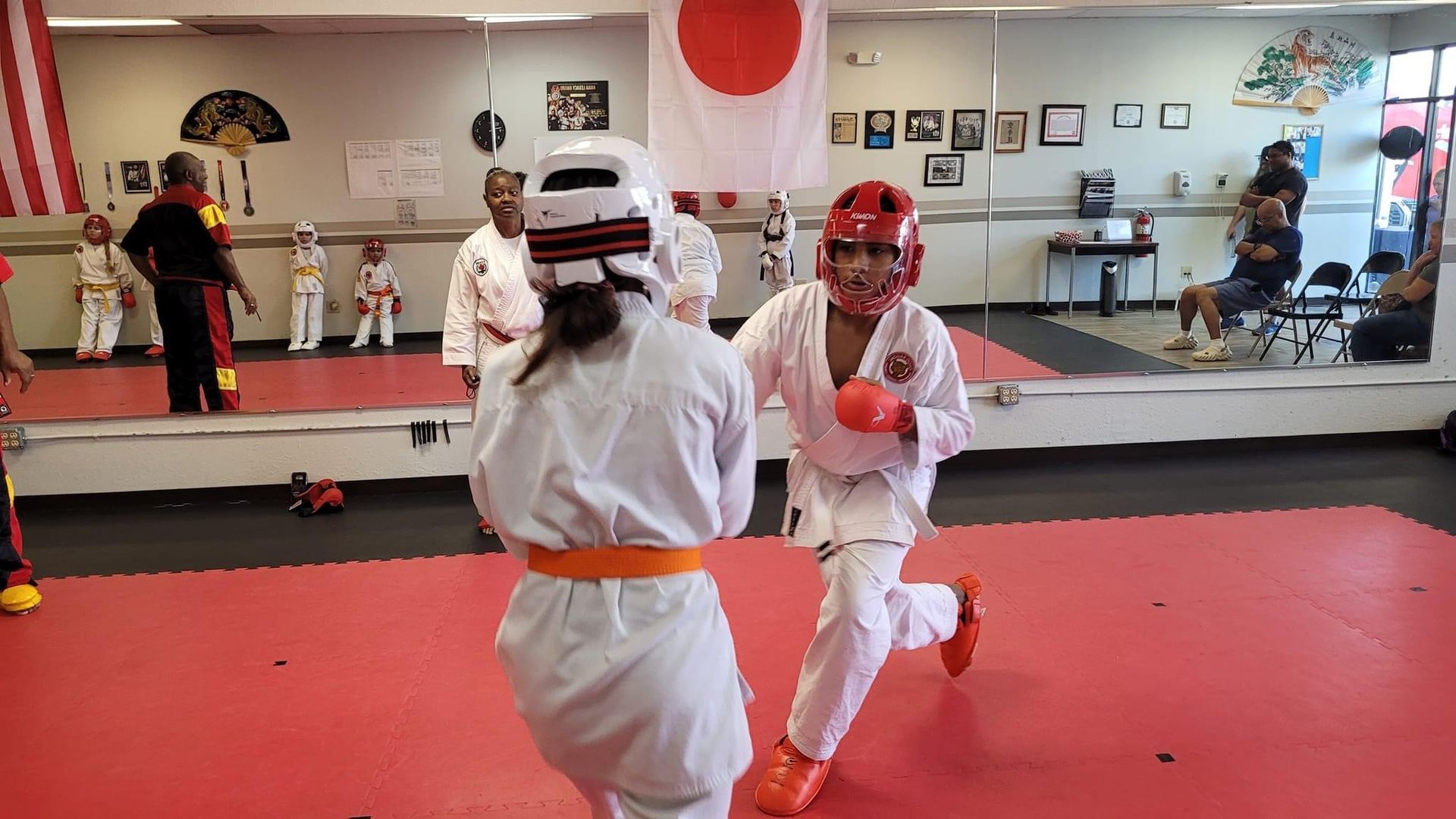 Two martial artists spar in a dojo, wearing white uniforms and protective gear on a red mat.