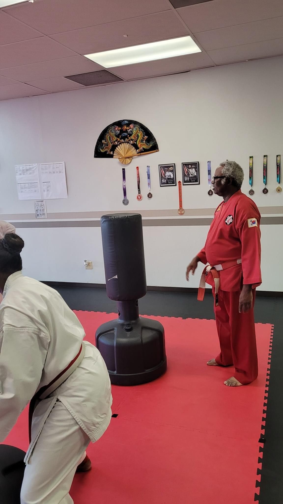 Martial arts class in a studio, with a person in a red uniform standing beside a black training dummy.