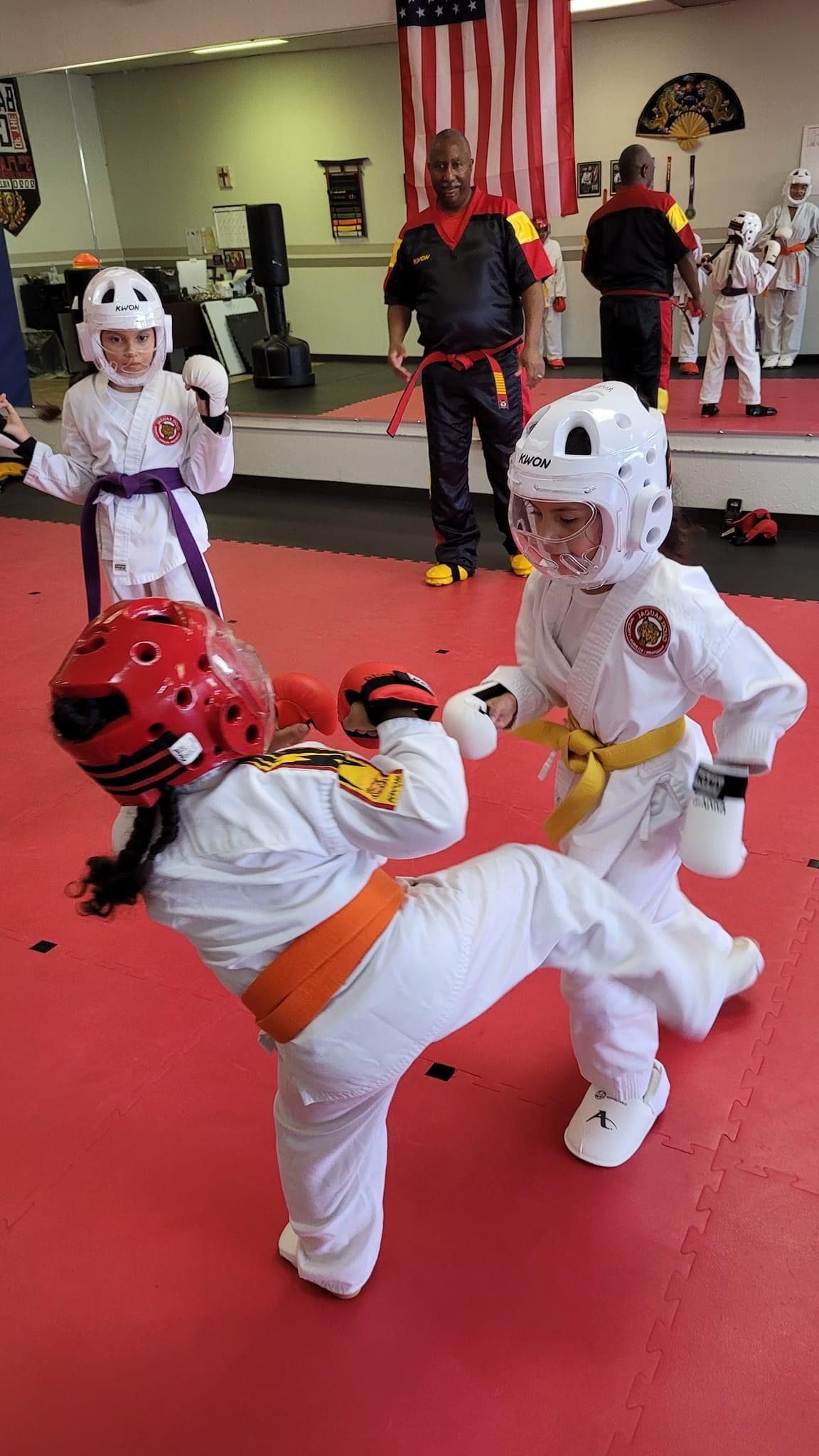 Two children sparring in white and red martial arts gear on a red mat in a dojo