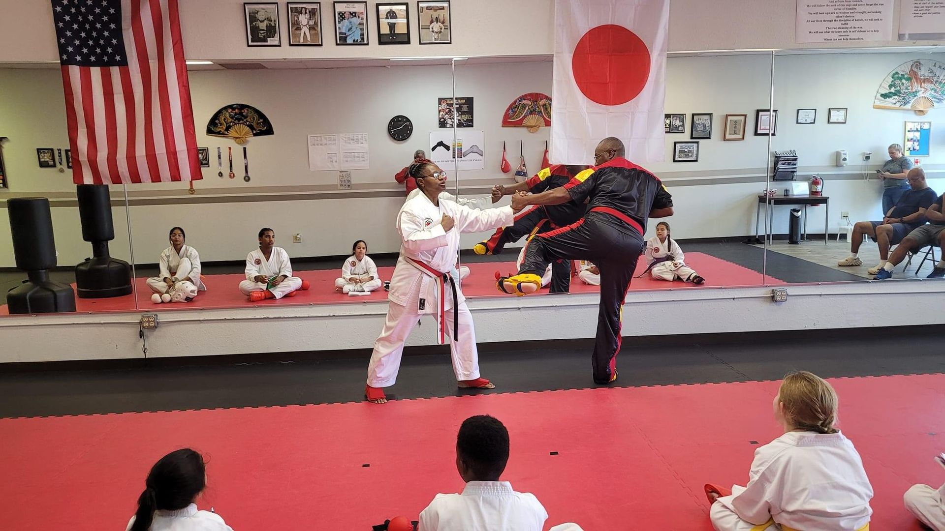 Two martial artists spar in a dojo, one kicking the other, with students seated behind them.