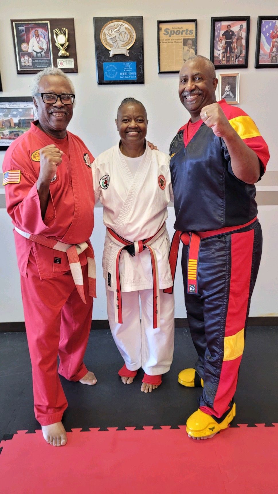 Three people in martial arts uniforms pose smiling in a dojo with framed awards on the wall.