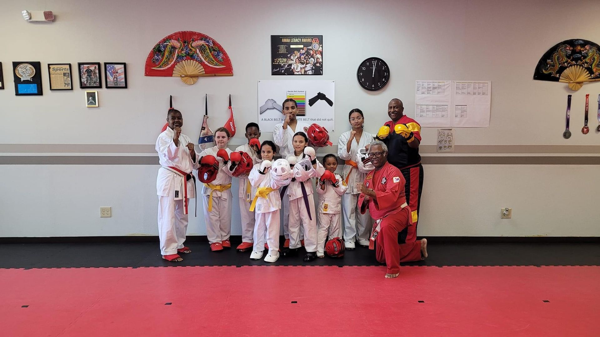 Group of martial arts students in white and red uniforms posing in a dojo with trophies on the wall.