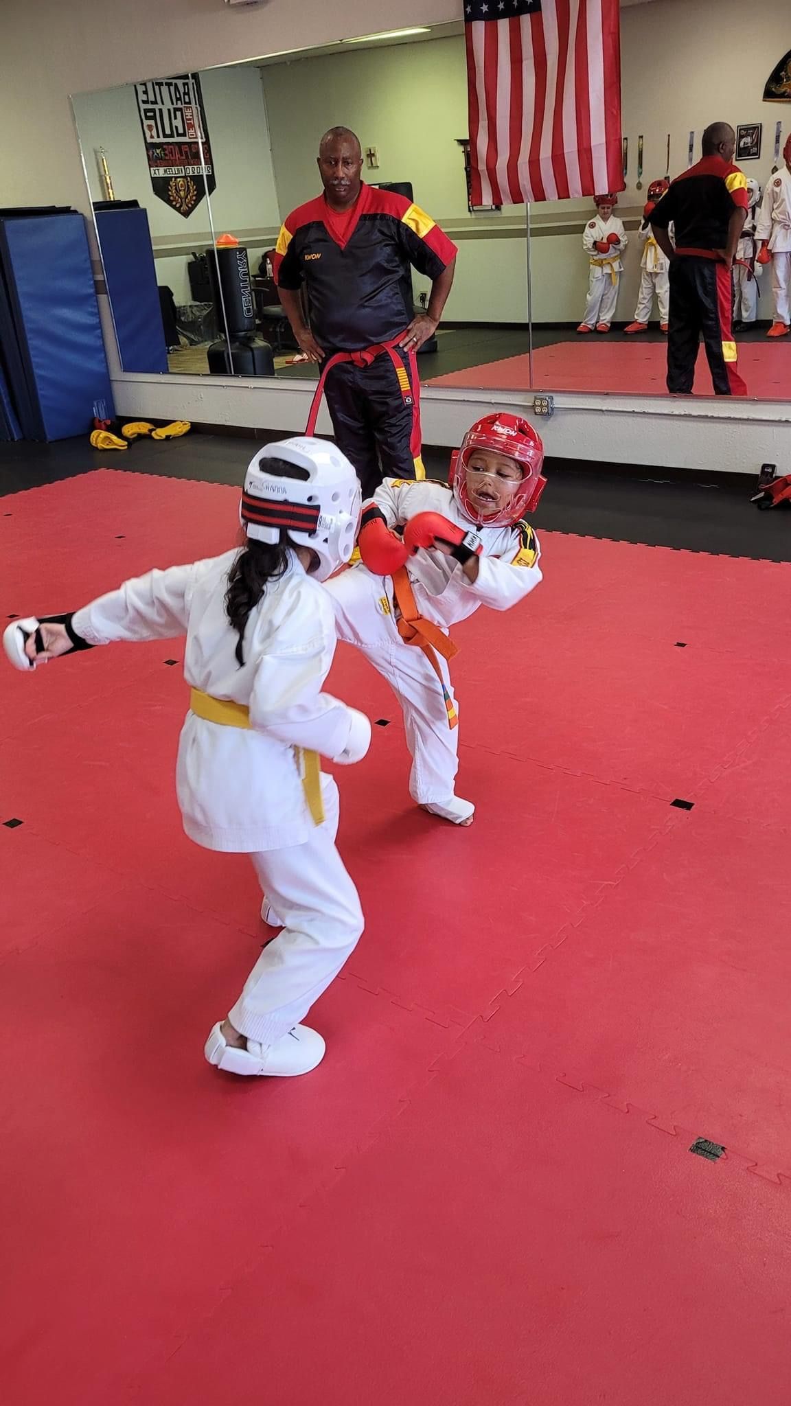 Two children in white martial arts uniforms sparring on red mats with an instructor in the background.