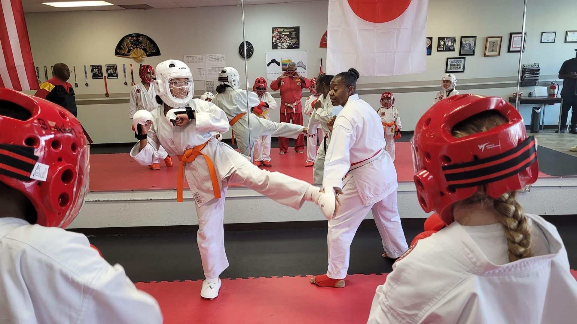 Karate class practicing a kick in a mirrored studio, with students in white uniforms and red sparring gear.