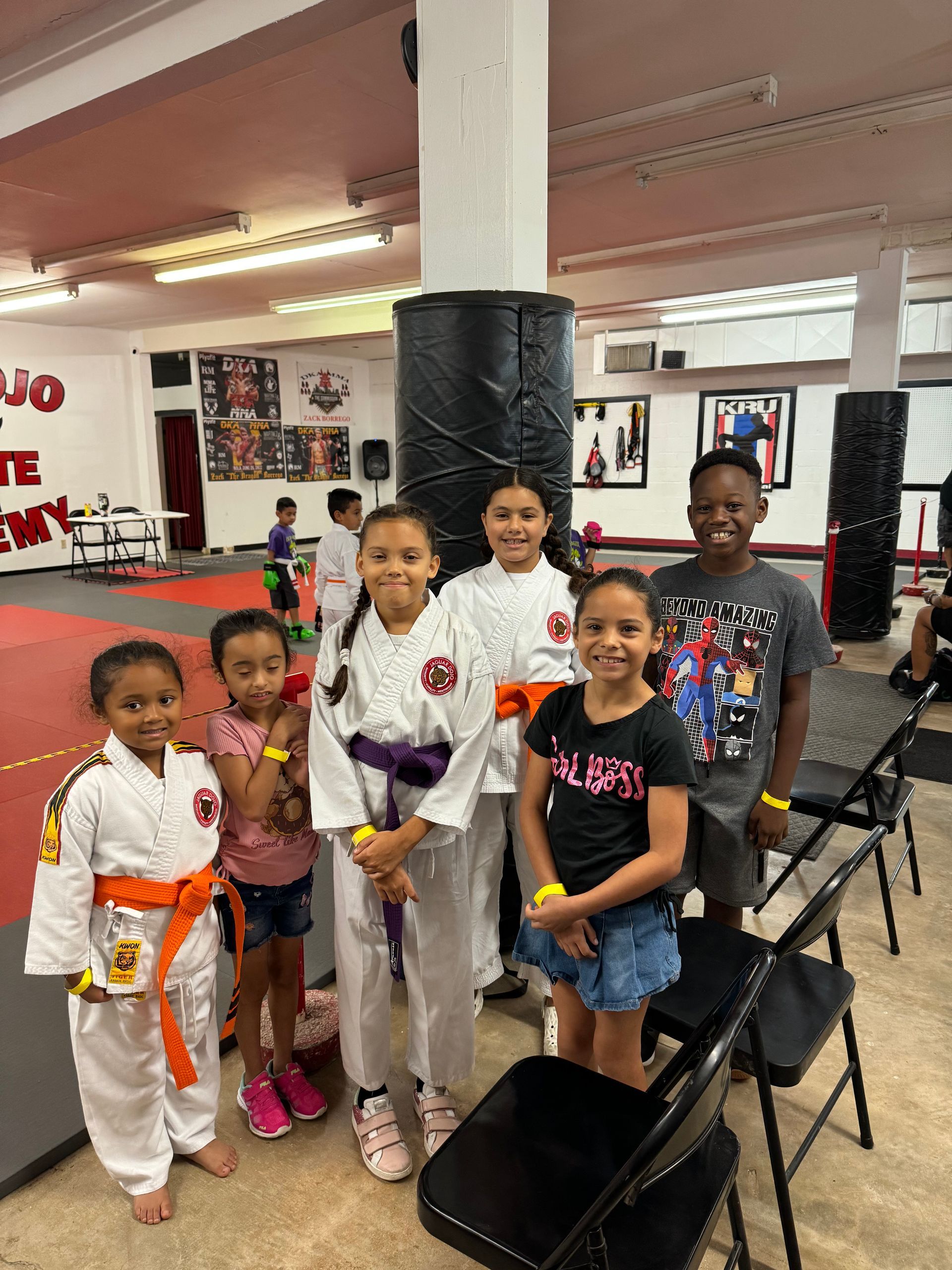 Six children posing in a gym with red mats, martial arts belts, and folding chairs.