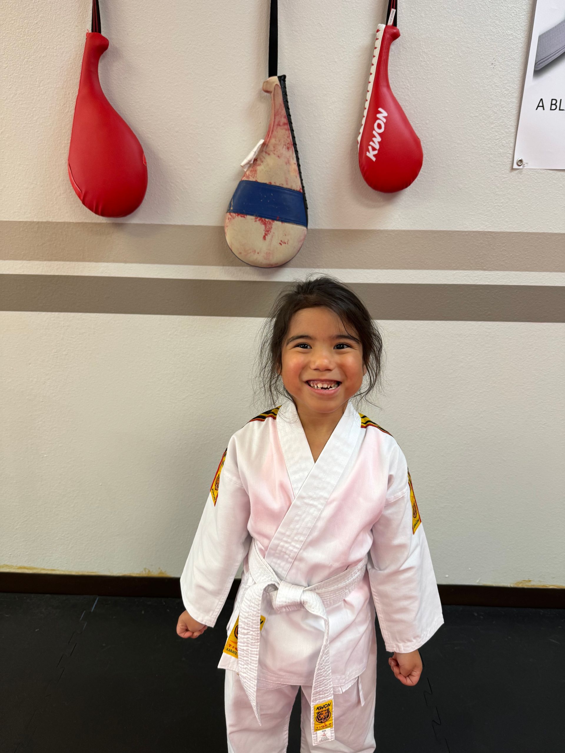 Smiling child in a white martial arts gi standing indoors beneath red and tan training bags.