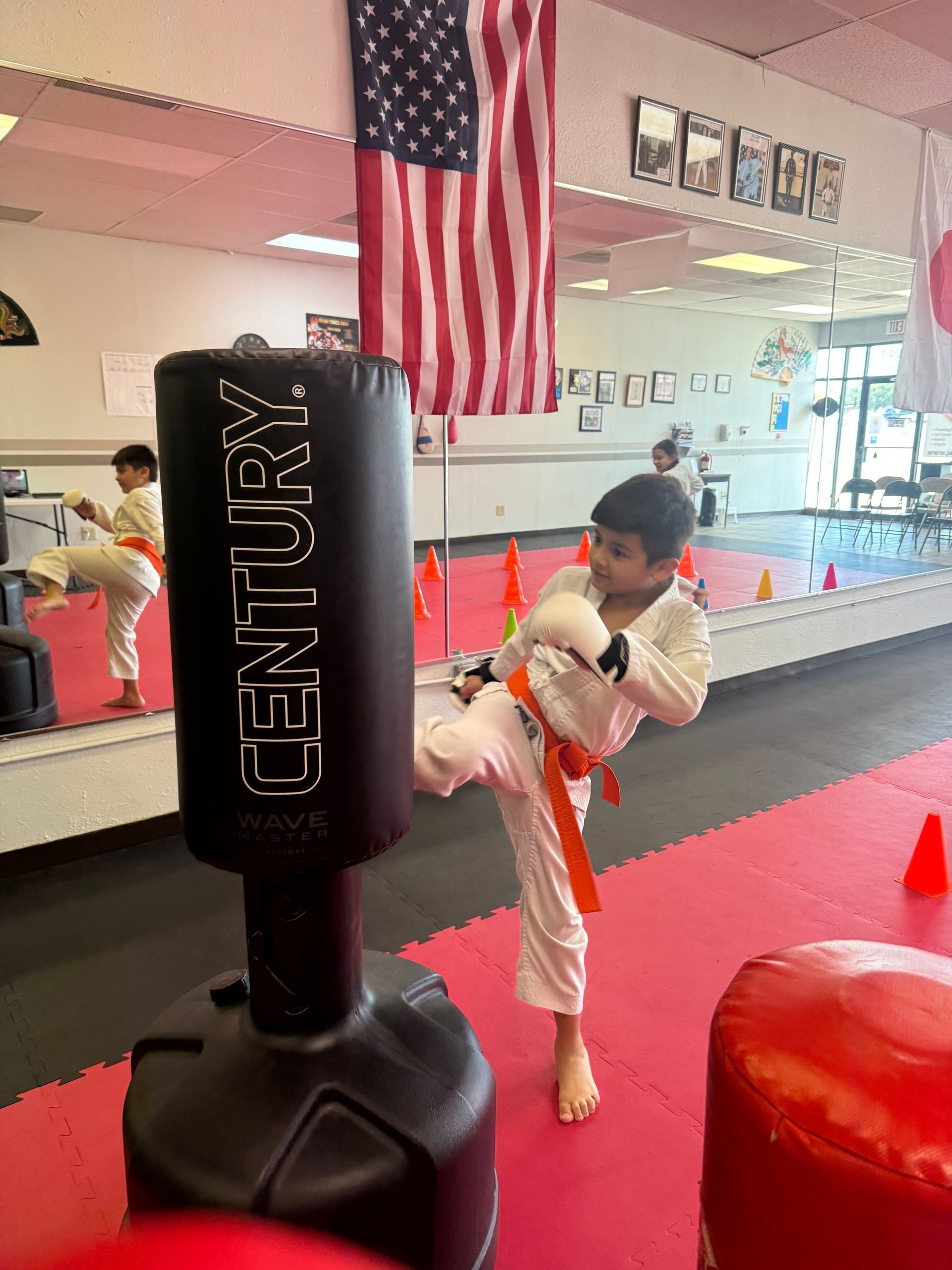 Child practicing a kick on a black punching bag in a martial arts studio with red mats and an American flag.