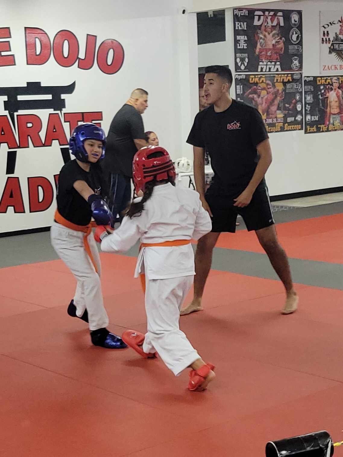 Two kids sparring in a martial arts dojo while two instructors watch nearby