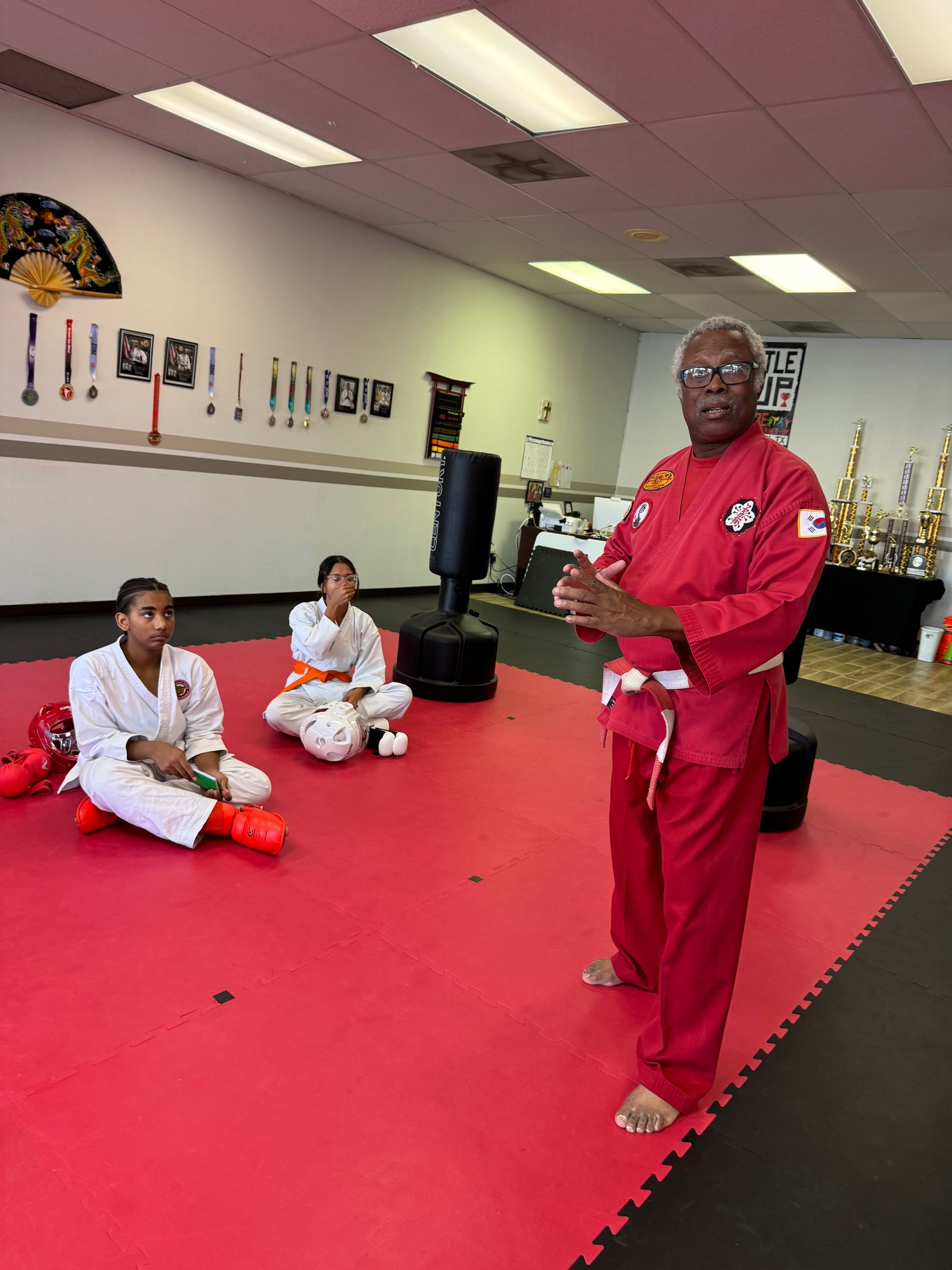 Martial arts instructor in red uniform speaking to two seated students in a dojo with red mats