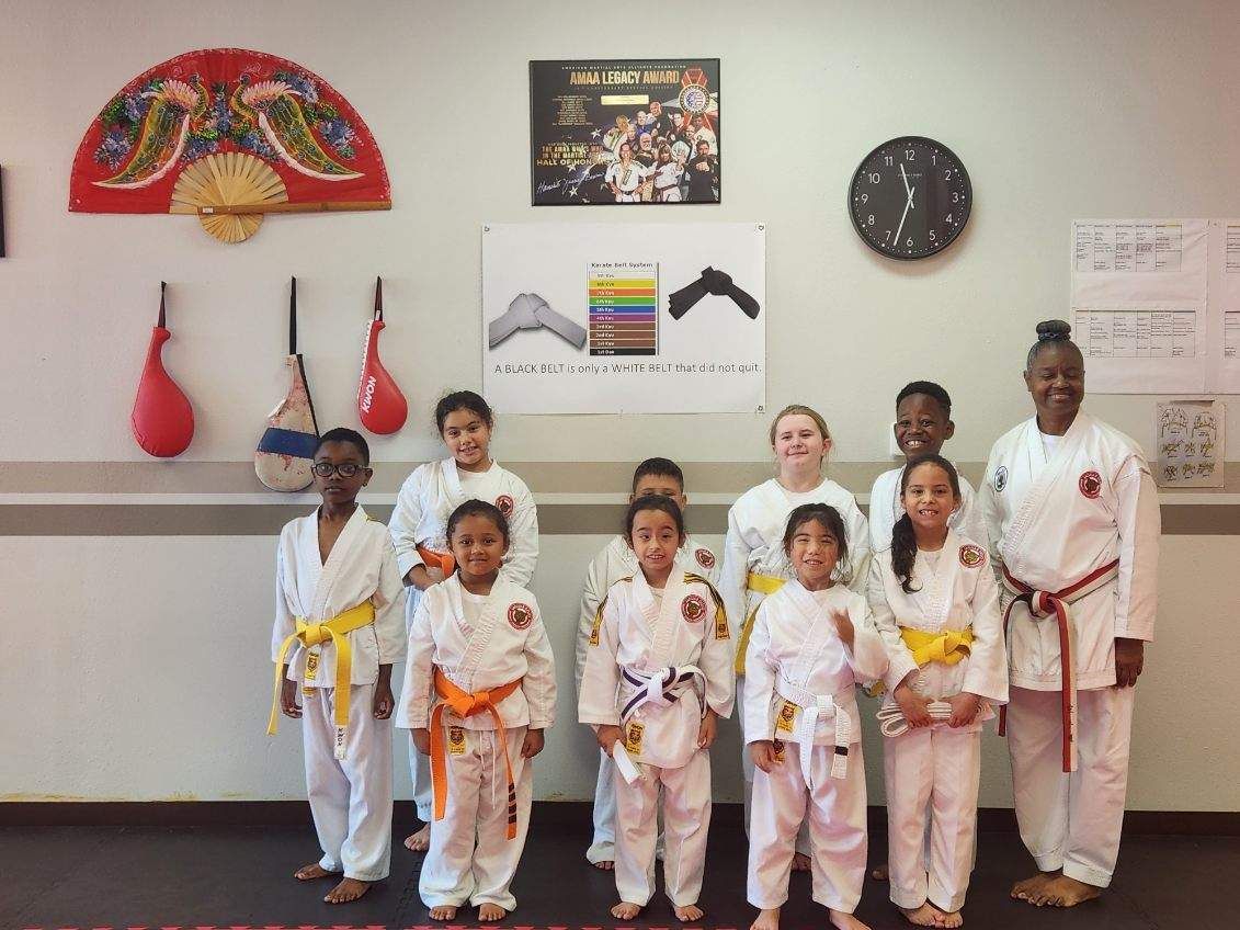 Group of children in martial arts uniforms posing with instructors in a dojo, holding yellow belts.