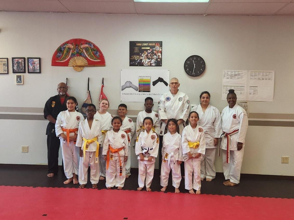 Children in white karate uniforms posing in a dojo with red mats and flags on the wall