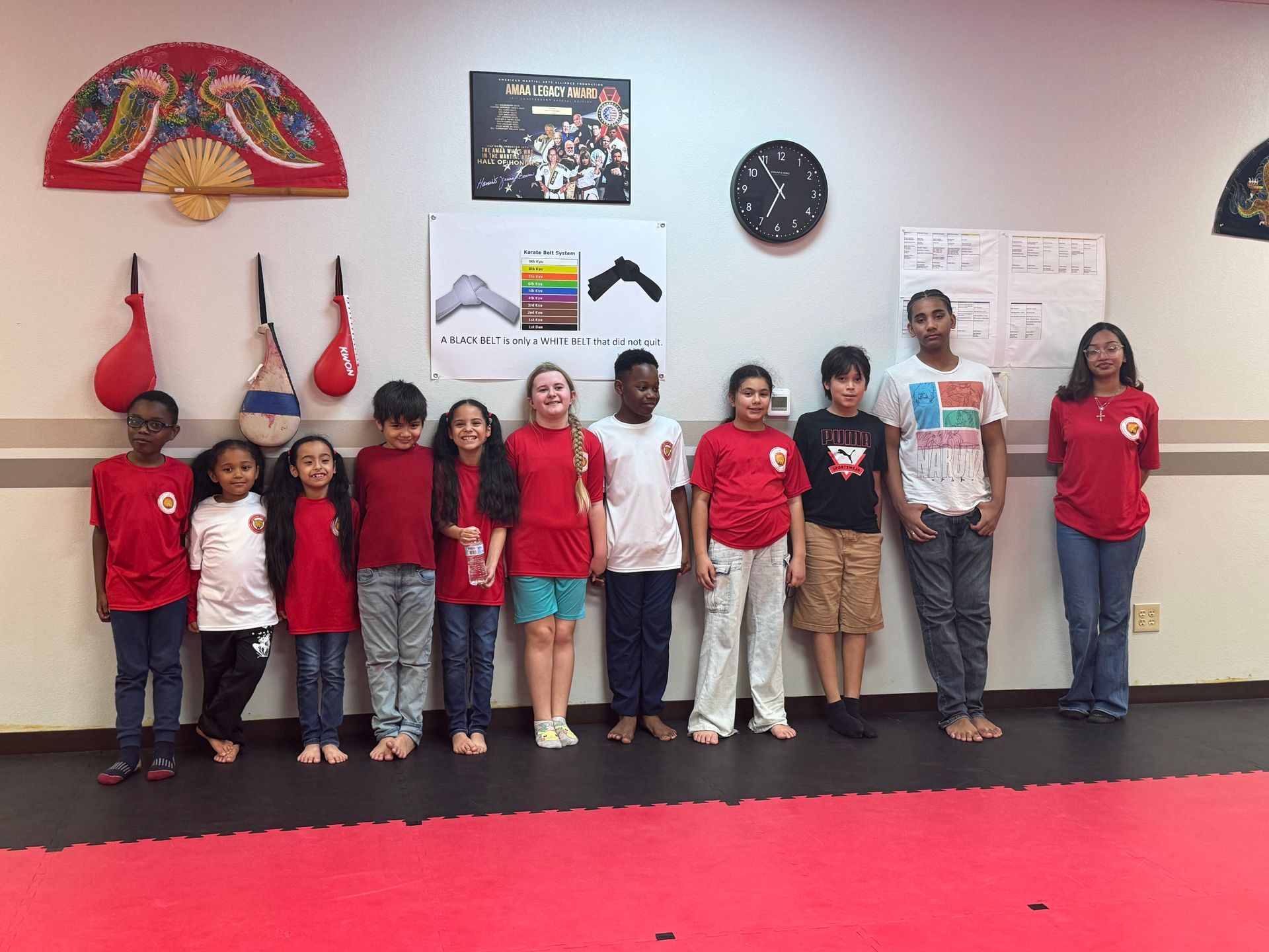 Group of children posing in red shirts against a gym wall with punching bags and martial arts signs