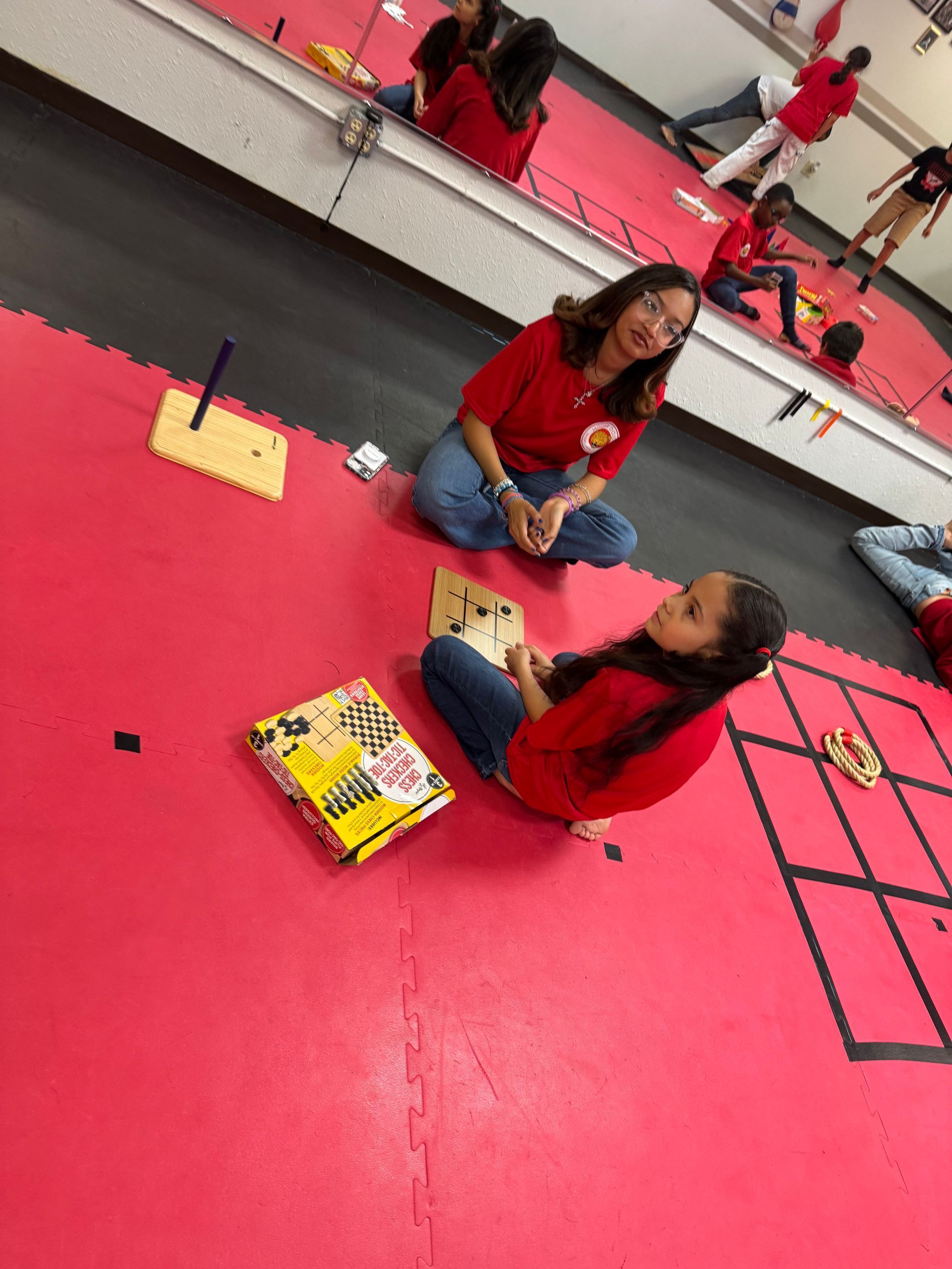 Two people sitting on a bright pink floor, facing each other in a mirrored room.
