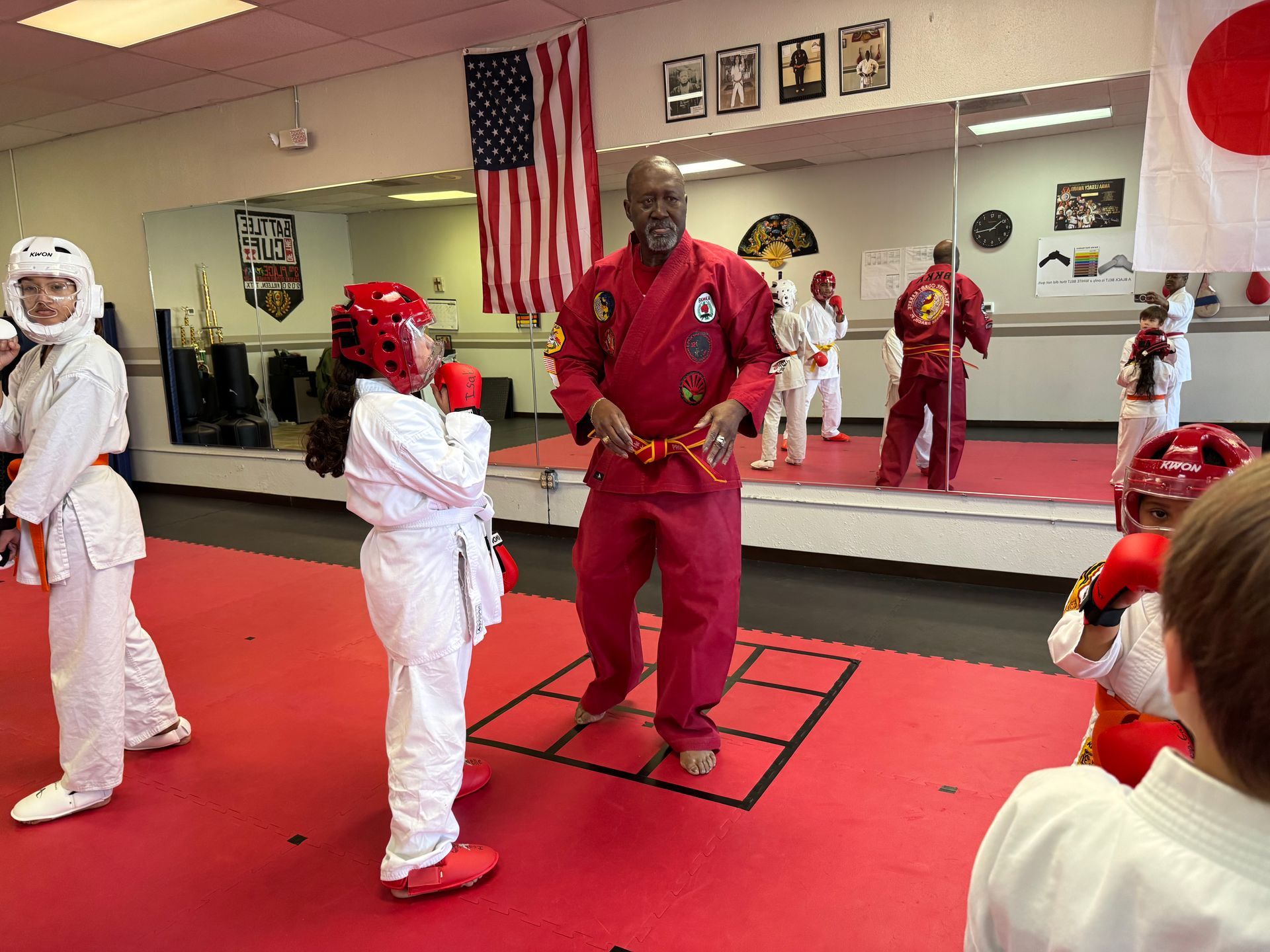 Martial arts class with students in white and red uniforms practicing in a mirrored studio on a red mat.
