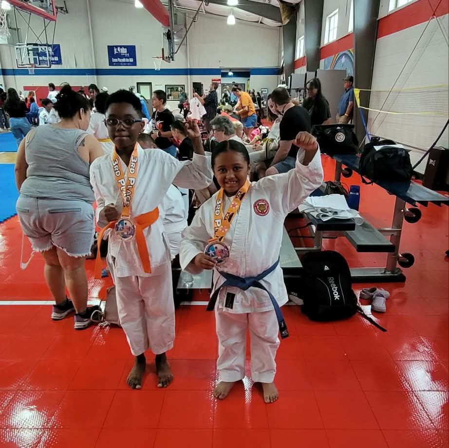 Two young martial artists in white uniforms pose with medals in a gym.