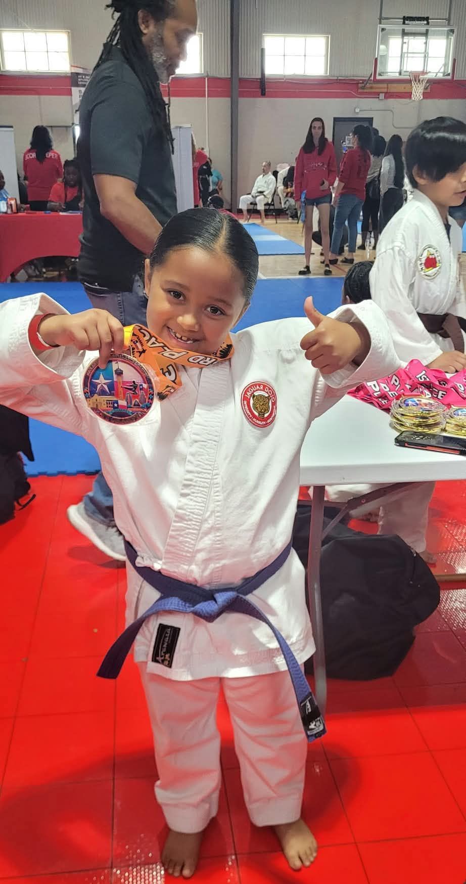Child in a white martial arts uniform holding a medal, posing in a red-floored gym after a competition.