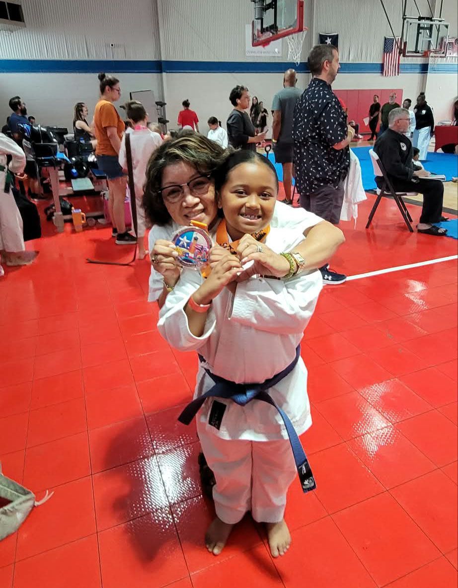 Two people in white martial arts uniforms smiling and holding medals in a gymnasium with a red mat.