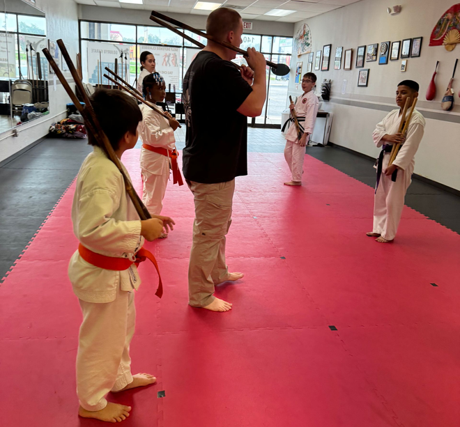 Martial arts class on red mats with students holding wooden swords while an instructor speaks.