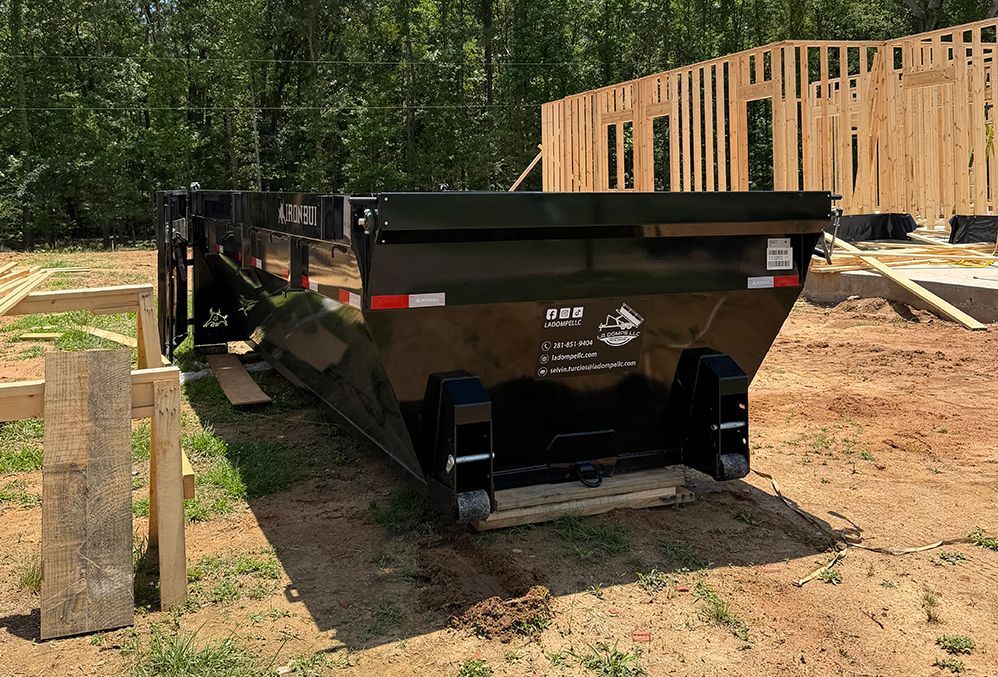 Black construction dumpster on dirt beside a wooden building frame in a wooded worksite