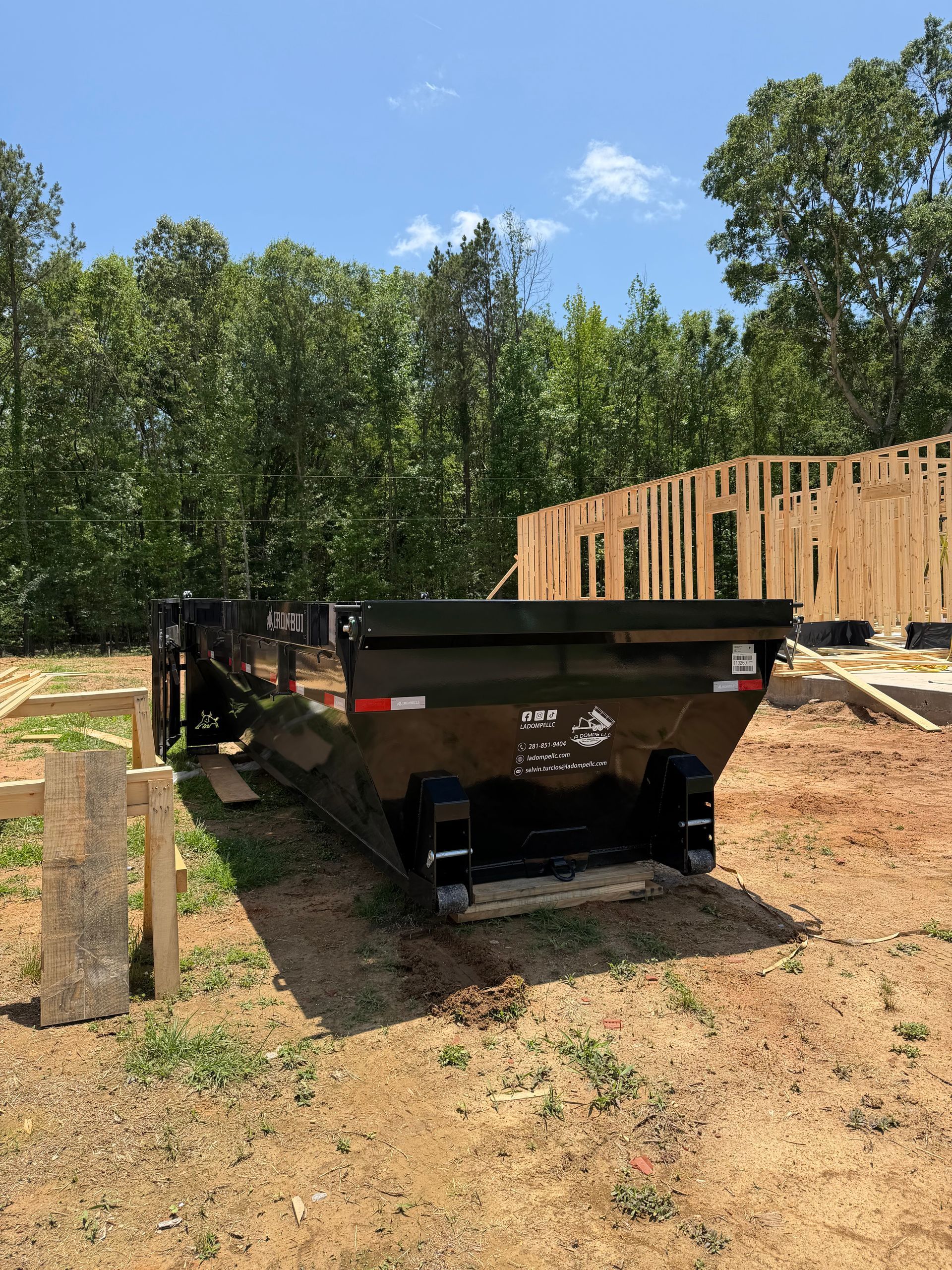 Black dump trailer at a construction site beside a wooden house frame and trees.