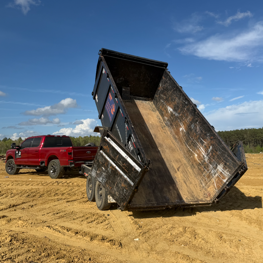 Red dump truck tipping a raised dump bed on a sandy dirt lot under a blue sky