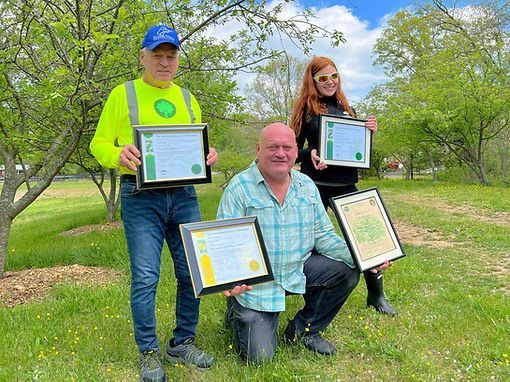 Three people kneel and stand outdoors in a park, holding framed certificates.