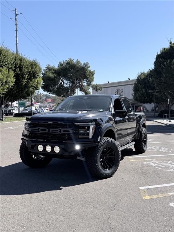 A black Ford Raptor truck with aftermarket lights and off-road tires parked in a sunny, paved parking lot.