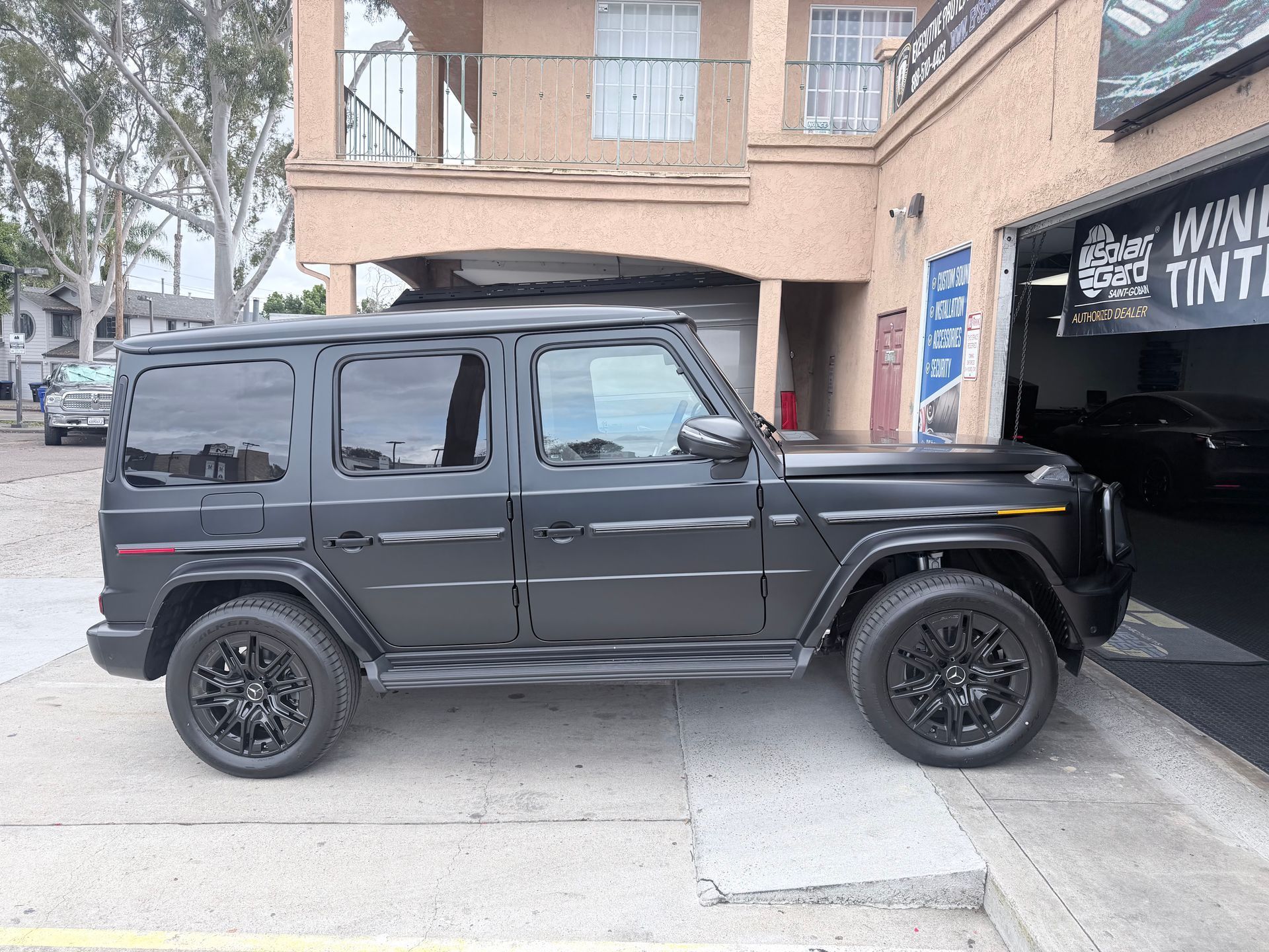A black Mercedes-Benz G-Class SUV parked on a concrete lot in front of a window tinting shop.