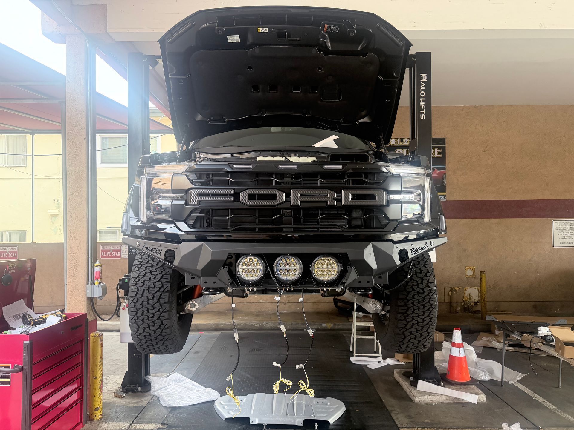 A black Ford Raptor truck raised on a hydraulic lift in a garage with its hood open and a skid plate removed.