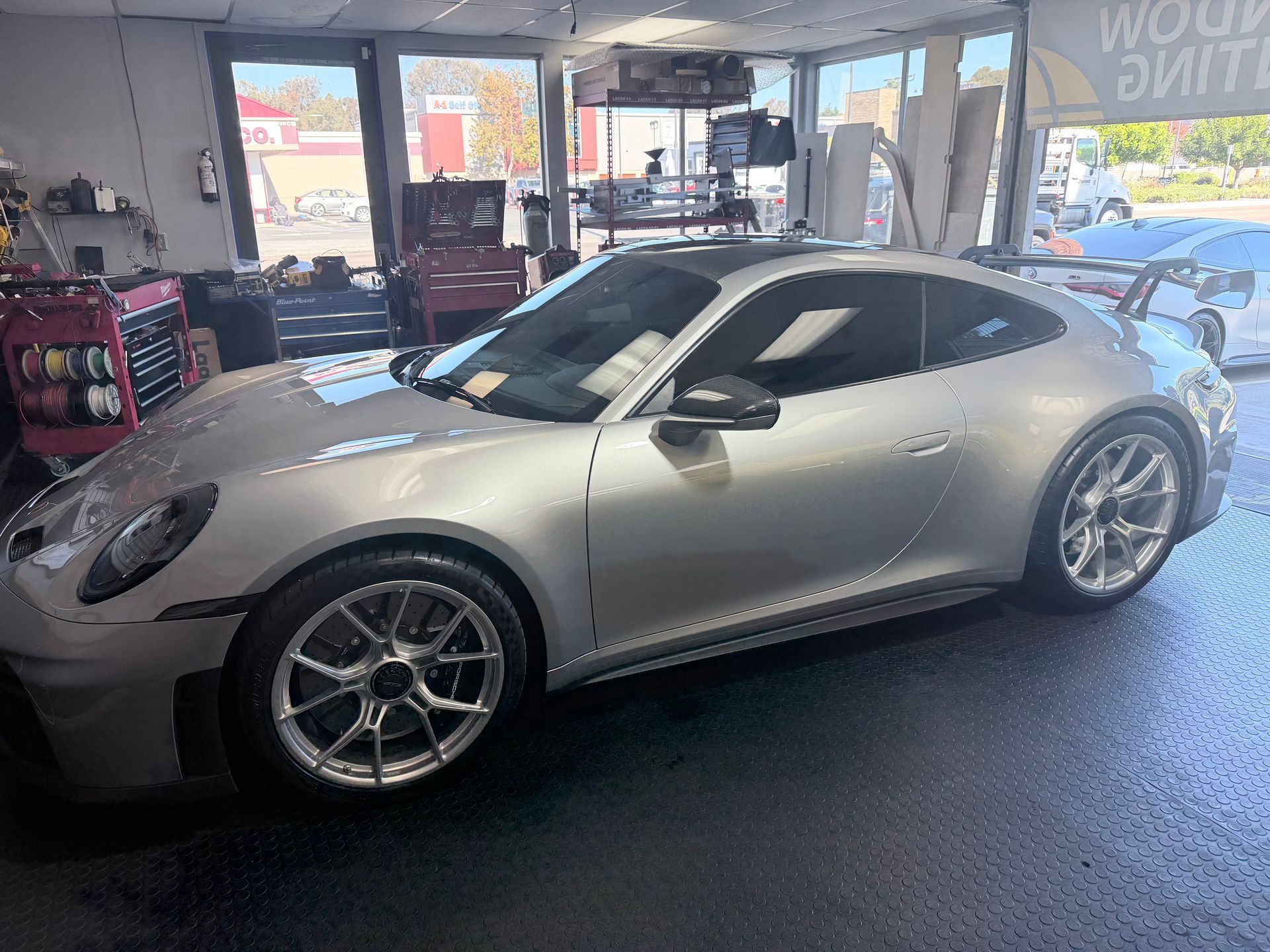 A silver Porsche parked inside an auto shop garage with bright, multi-spoke wheels.