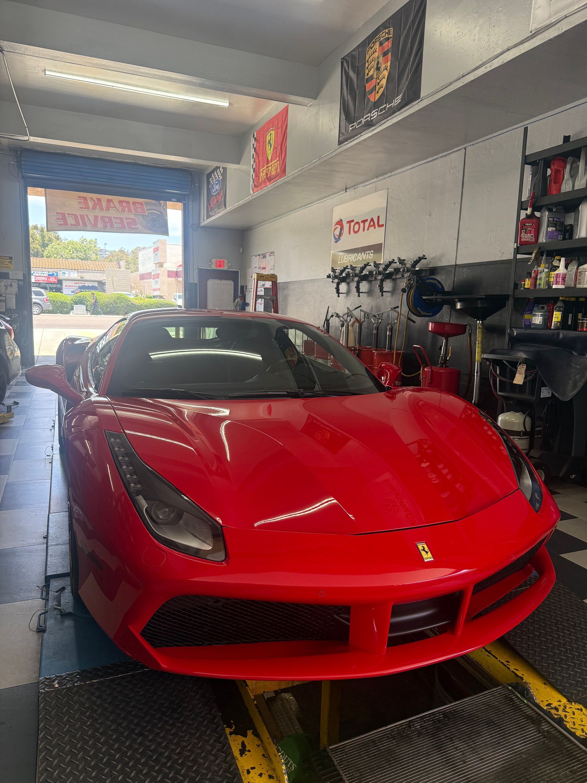 A red Ferrari parked inside an automotive repair shop with tools and shelves visible in the background.