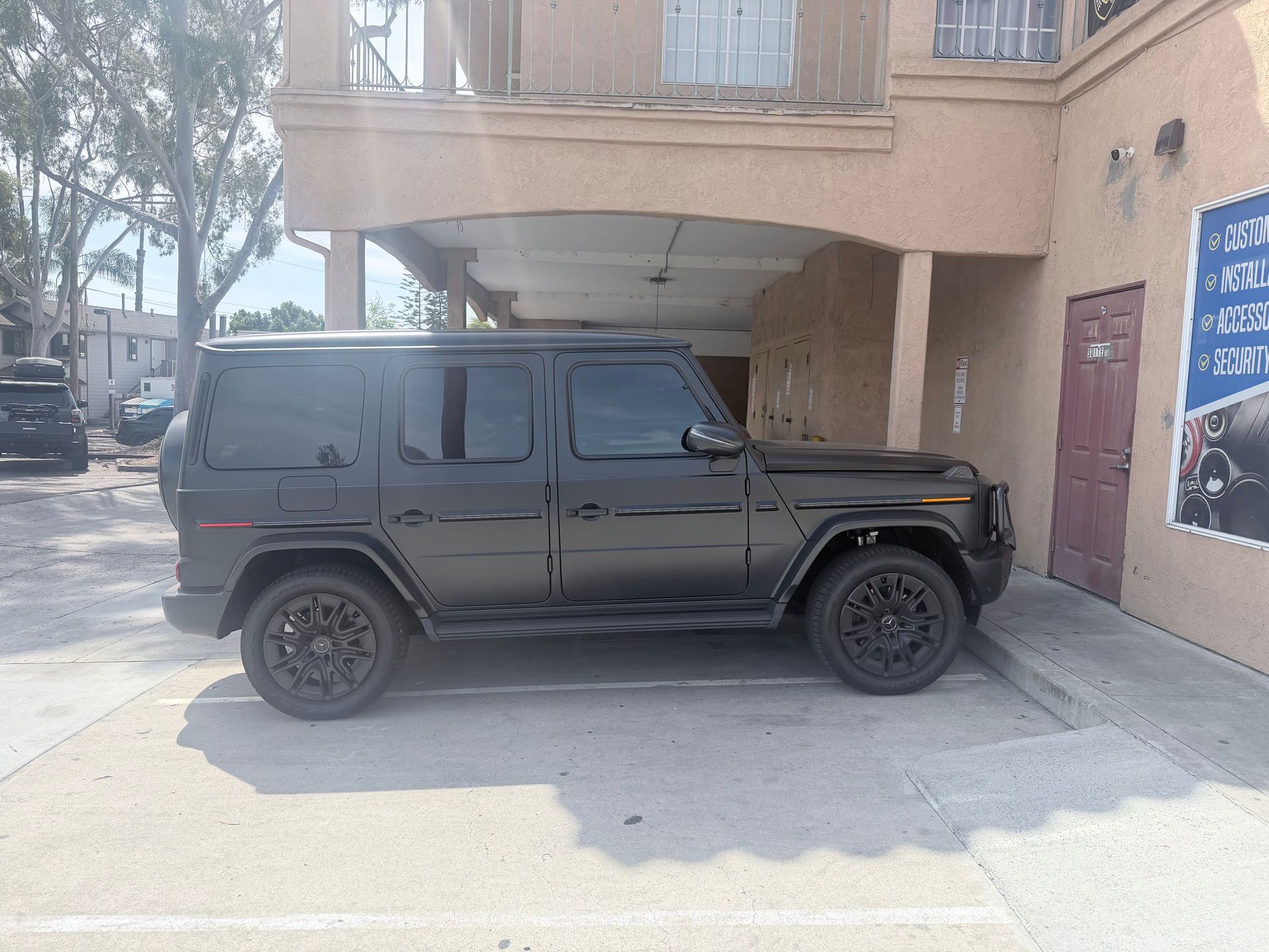 A black Mercedes-Benz G-Class SUV parked in a paved lot in front of a multi-story building.