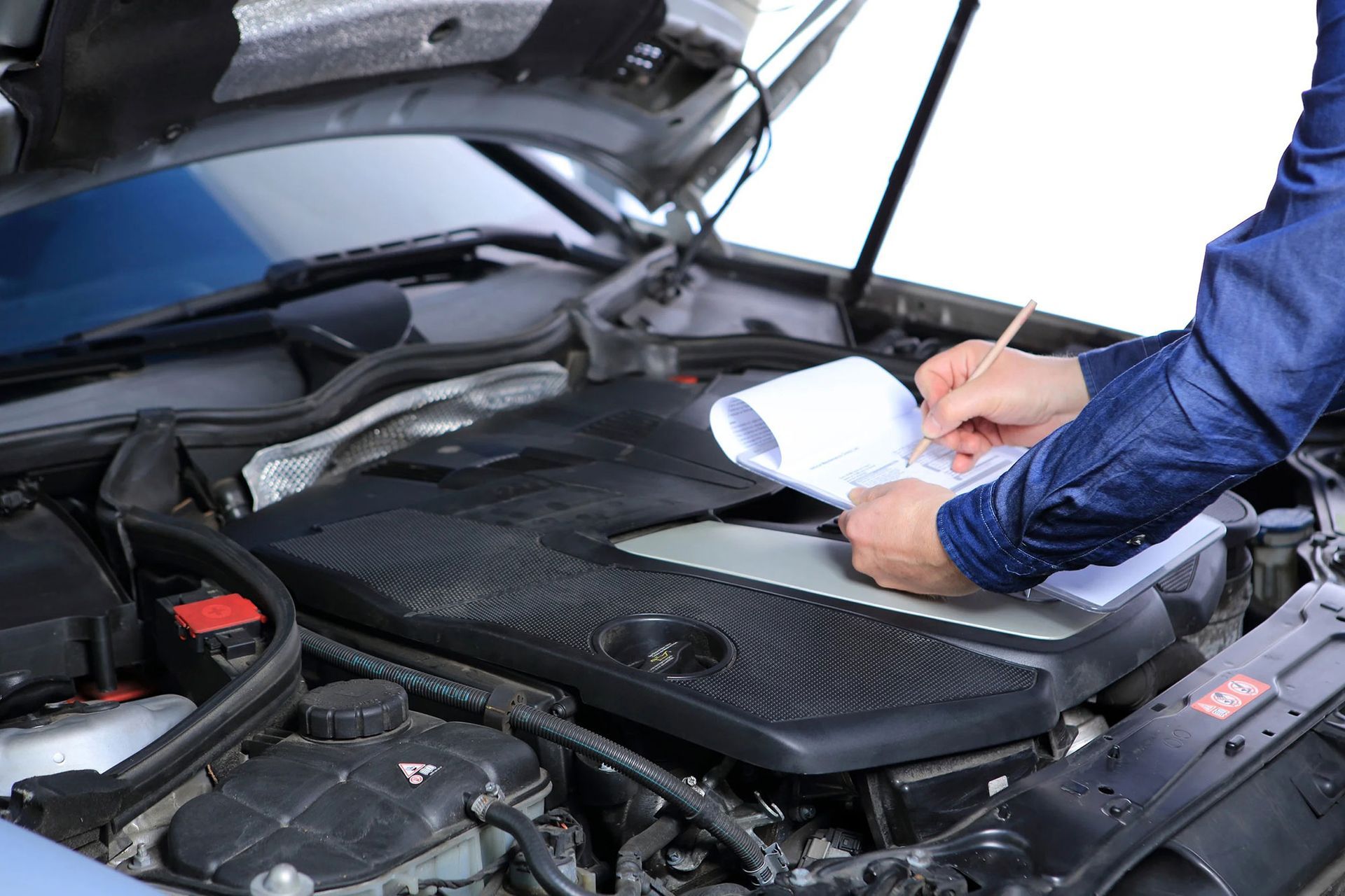 Mechanic inspects car engine, writing on a clipboard with the hood open.