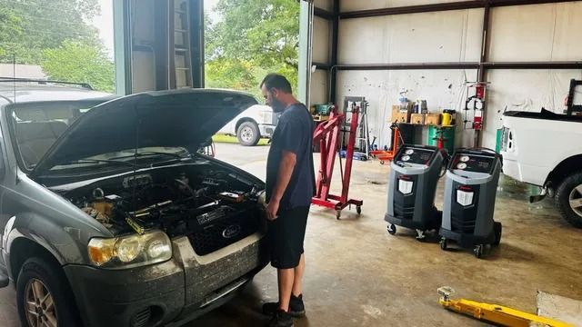 Mechanic examining a car engine in a garage; gray car, tools, equipment visible.