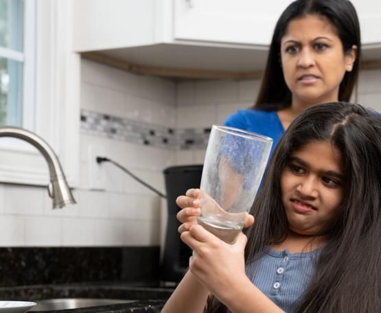 Girl holds up an empty glass in a kitchen while an adult stands behind her, looking concerned.