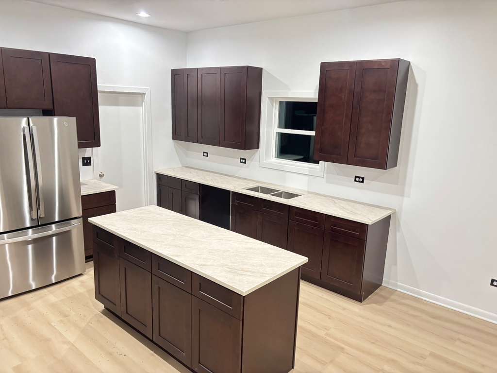 Empty kitchen with dark cabinets, stainless steel fridge, light countertops, and a central island.