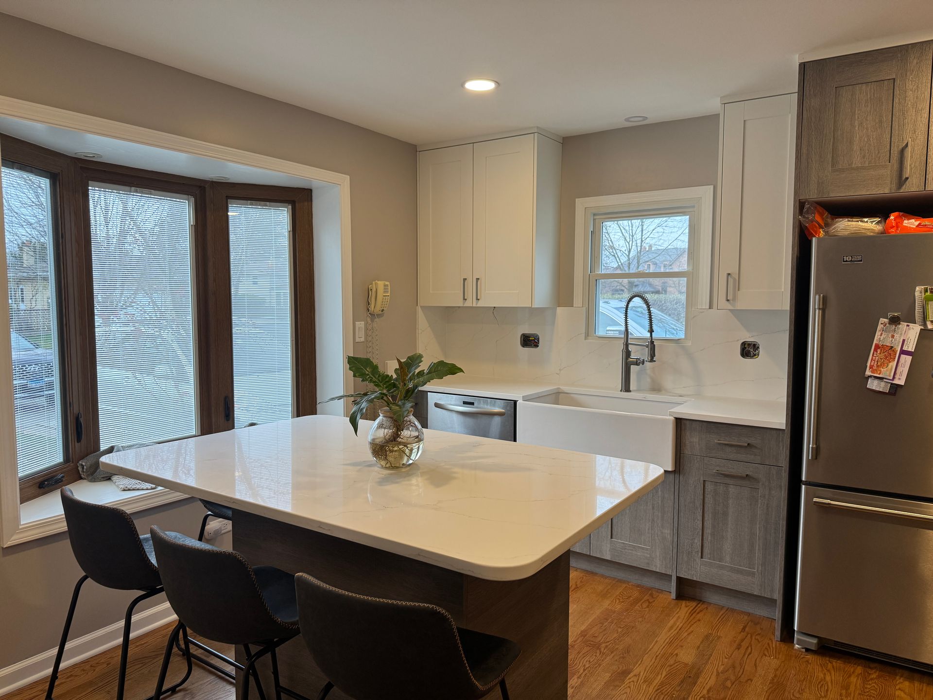 Modern kitchen with white island, black stools, wood cabinets, stainless fridge, and large windows
