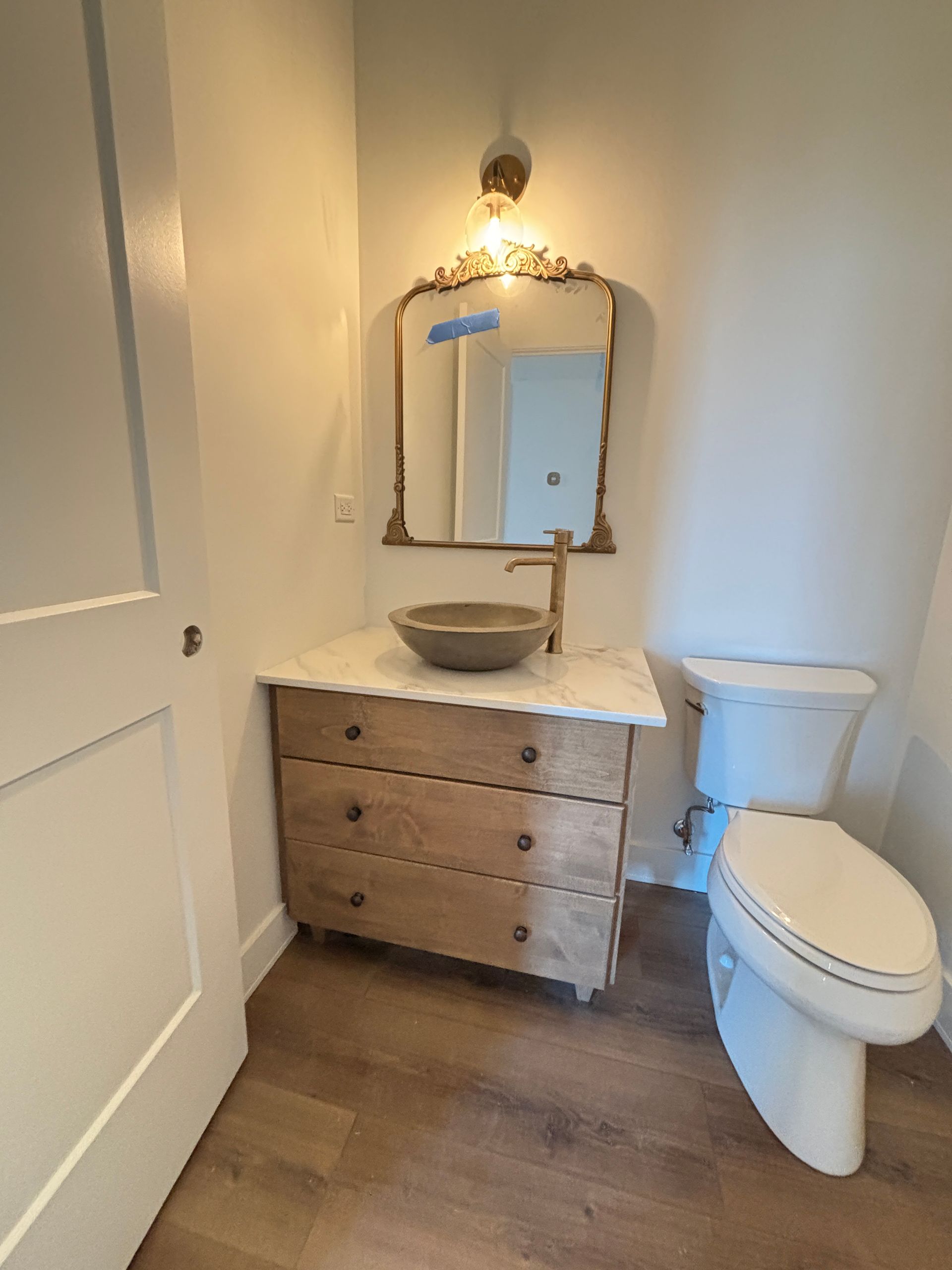 Small bathroom with a wood vanity, round mirror, vessel sink, toilet, and white walls.