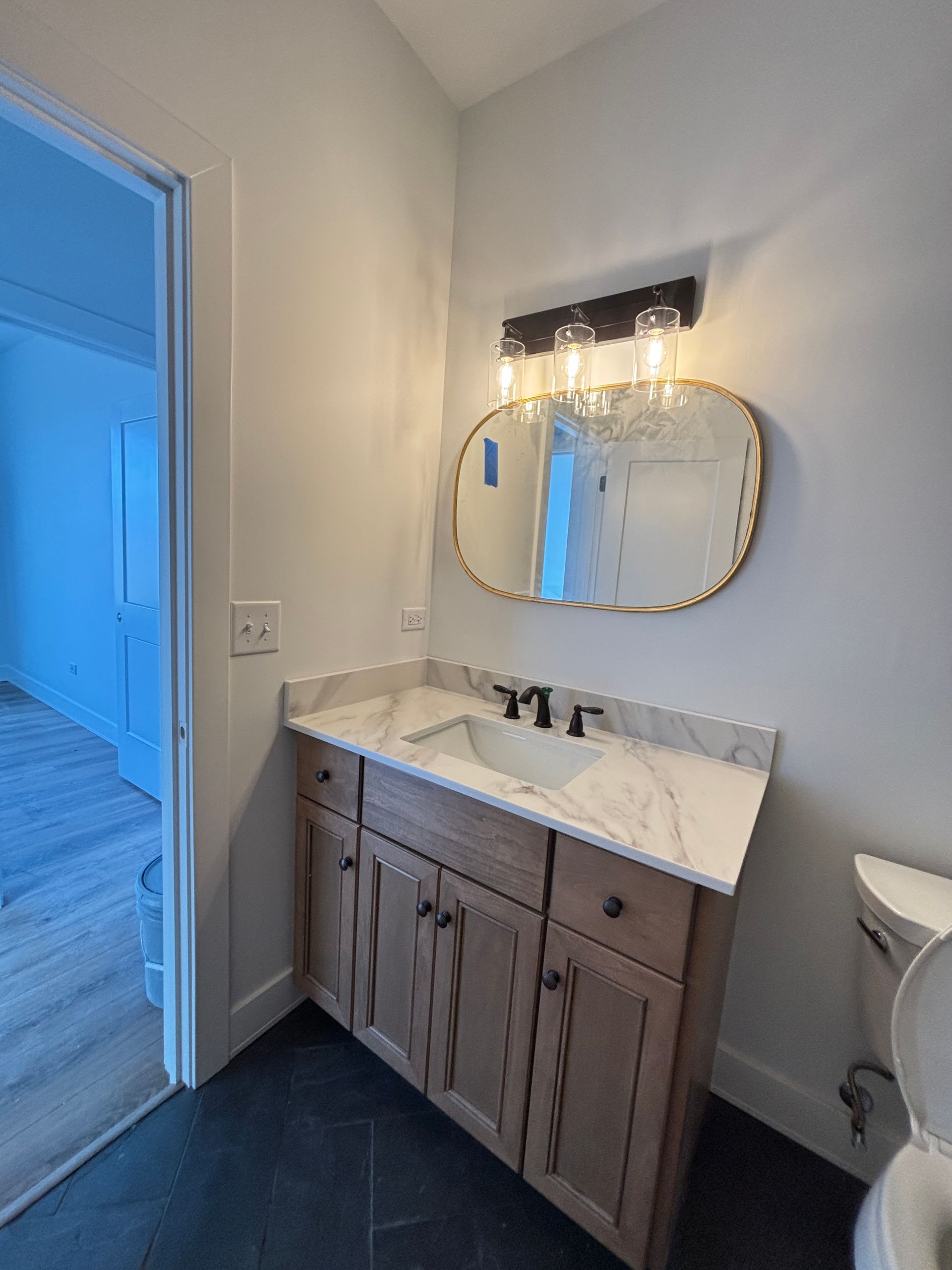 Small bathroom vanity with marble countertop, round mirror, and wall sconce beside a blue-lit doorway