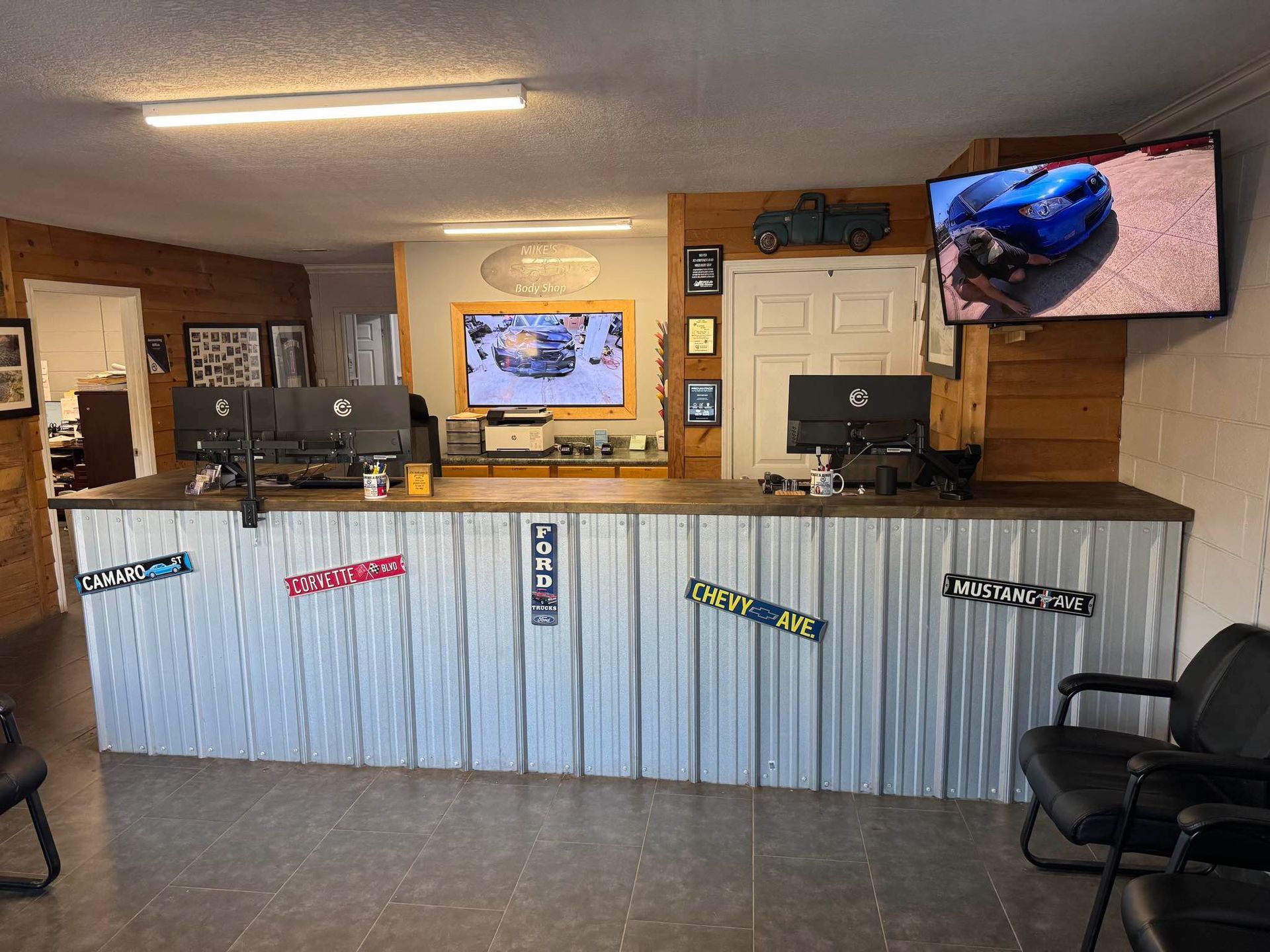 An office reception area with a corrugated metal front desk, computers, two wall-mounted televisions, and black seating.