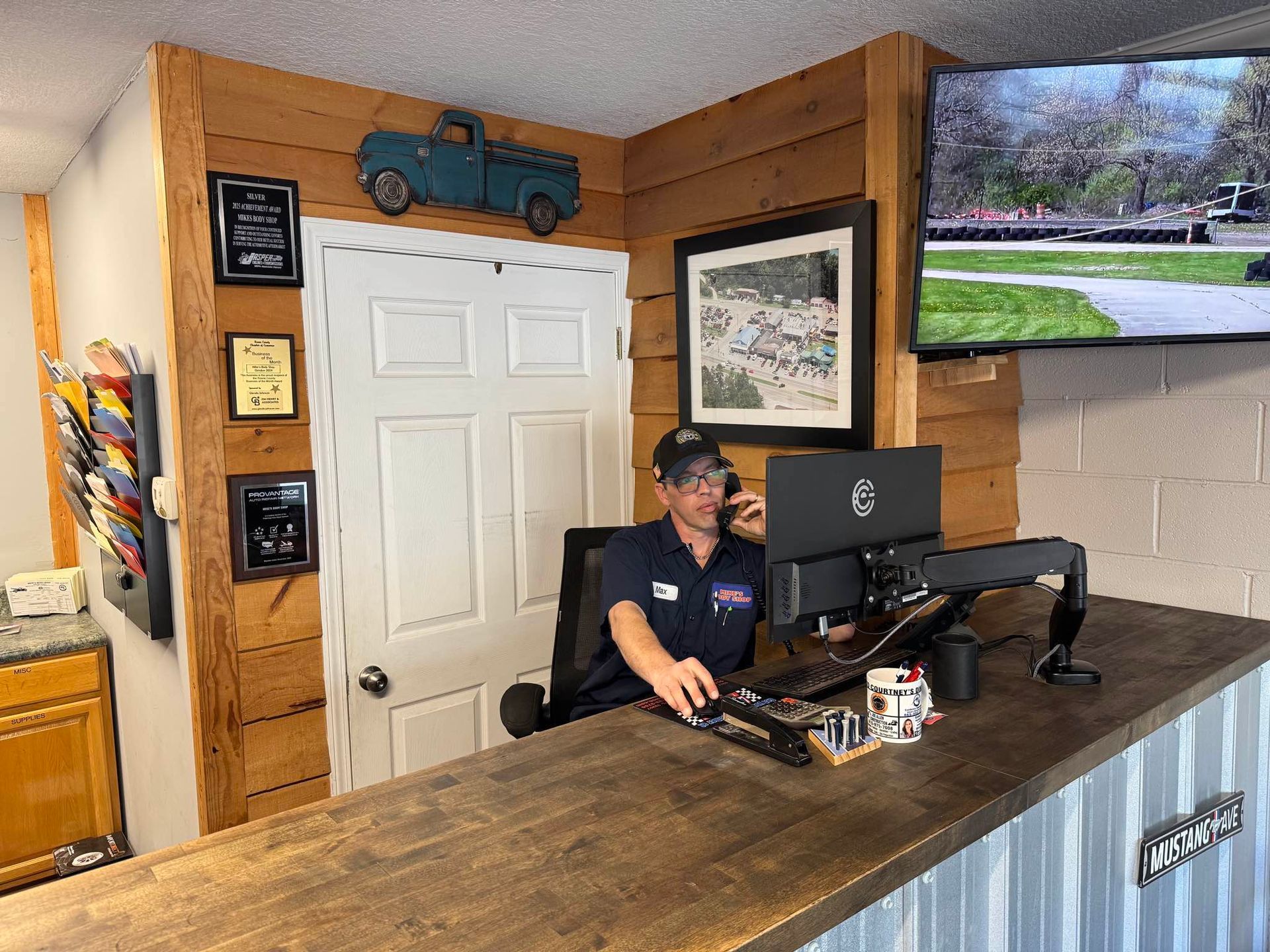 A person sitting at a wood-paneled office reception desk, working on a computer near a white door and wall-mounted decor.