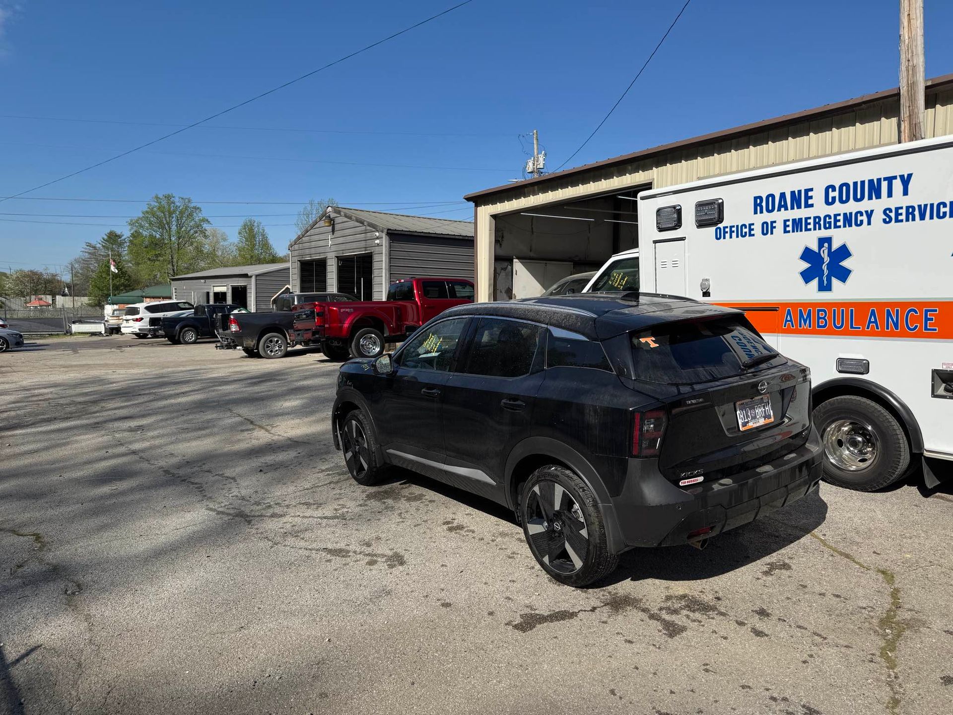 A black SUV parked next to a Roane County ambulance outside a garage building under a clear blue sky.